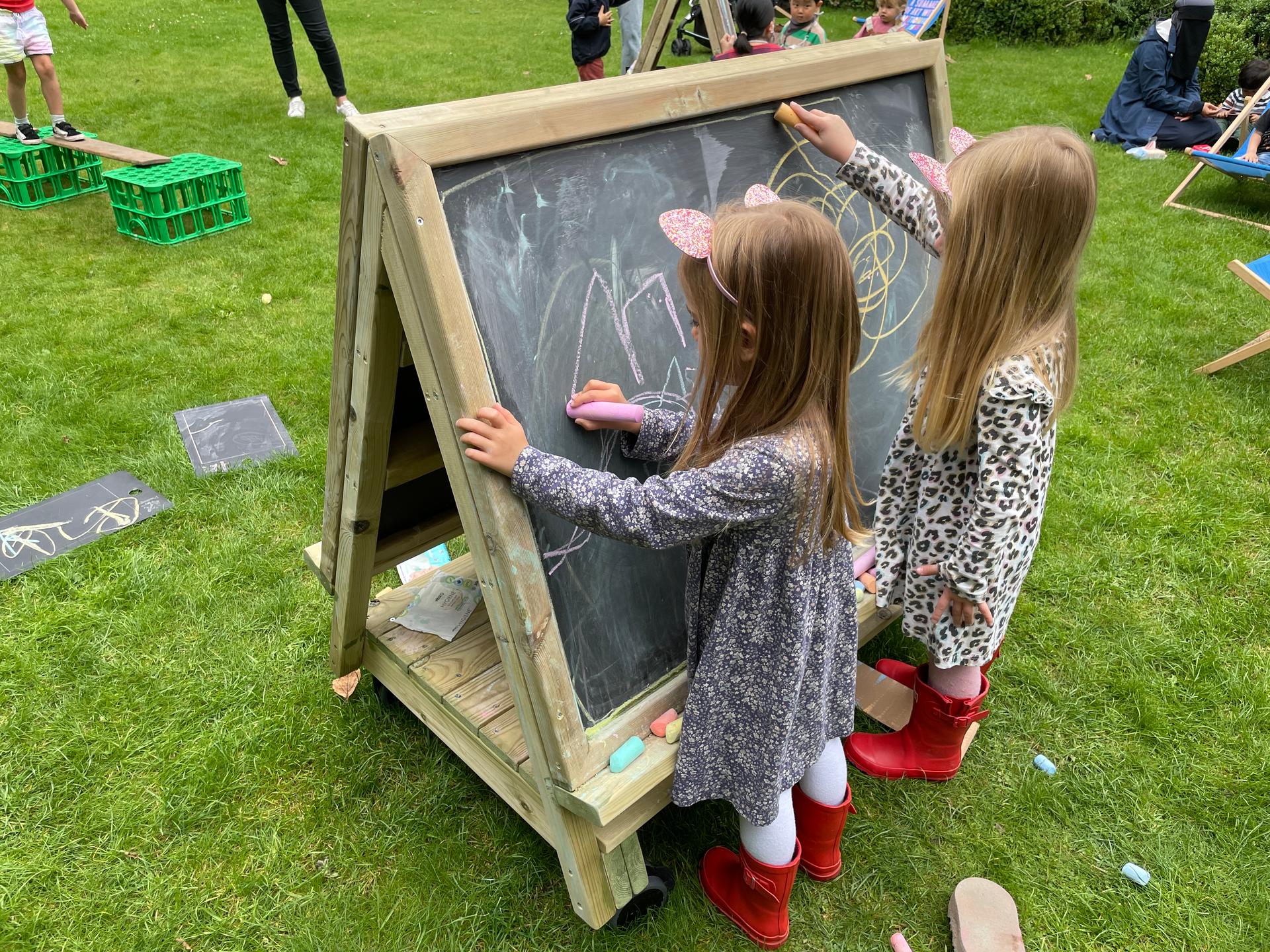 Children drawing on a wooden, chalkboard easel on wheels.
