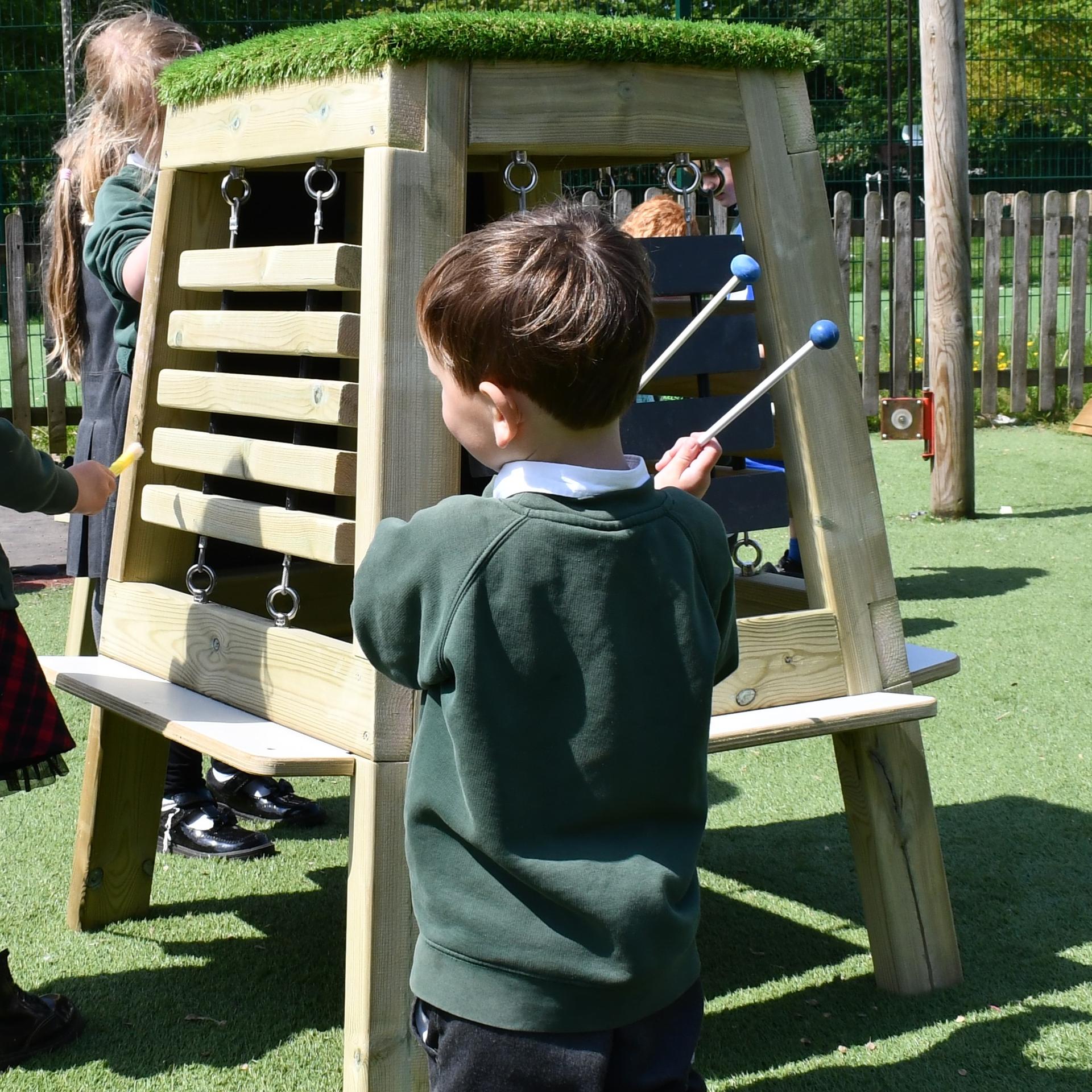 A little boy is kneeling beside the Group Music Easel as he holds two beaters over his right shoulder.