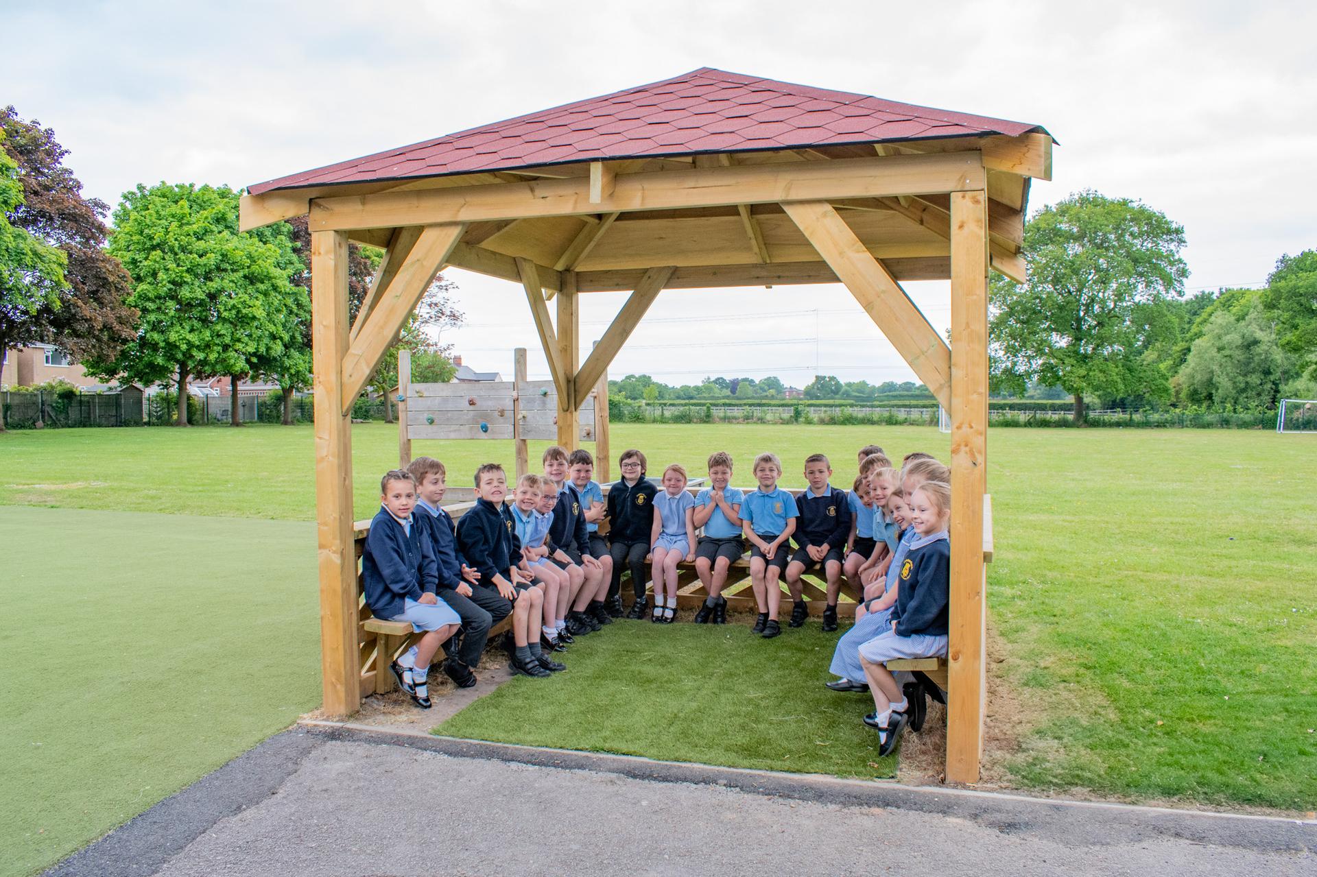 A group of school children in blue uniform, sitting on wooden benches within a in wooden 3m gazebo, pitched on grass, outside a school playing field.