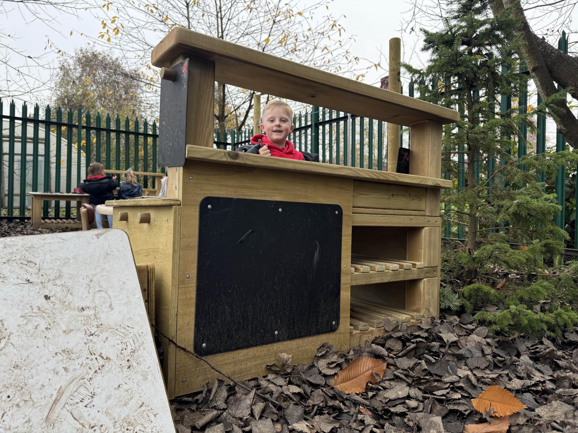 A boy is nursery is looking over the Wooden Mud Kitchen towards the camera.