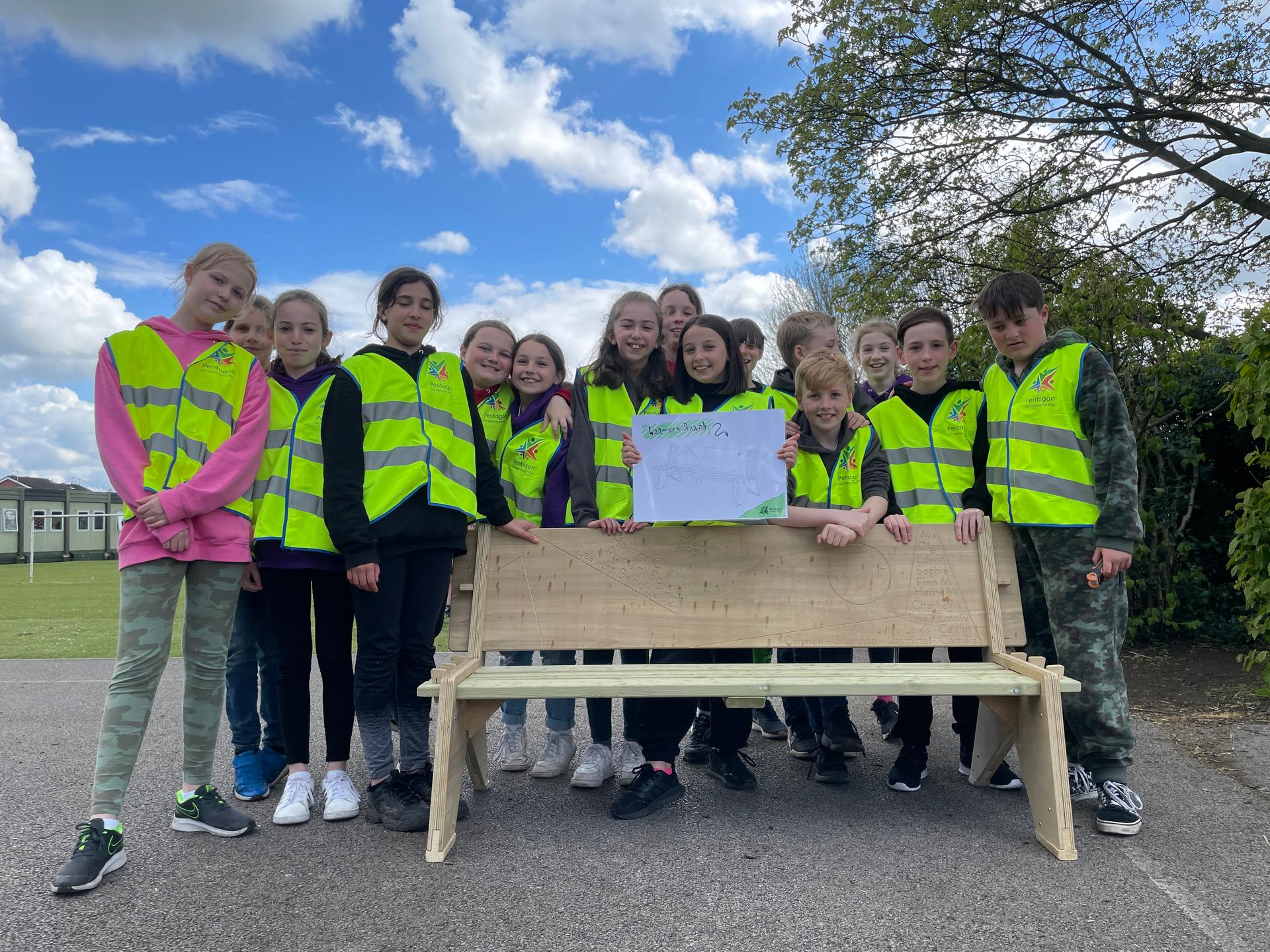 A group of Y6 children standing next to a recently built leavers bench with all of their names displayed on the back.