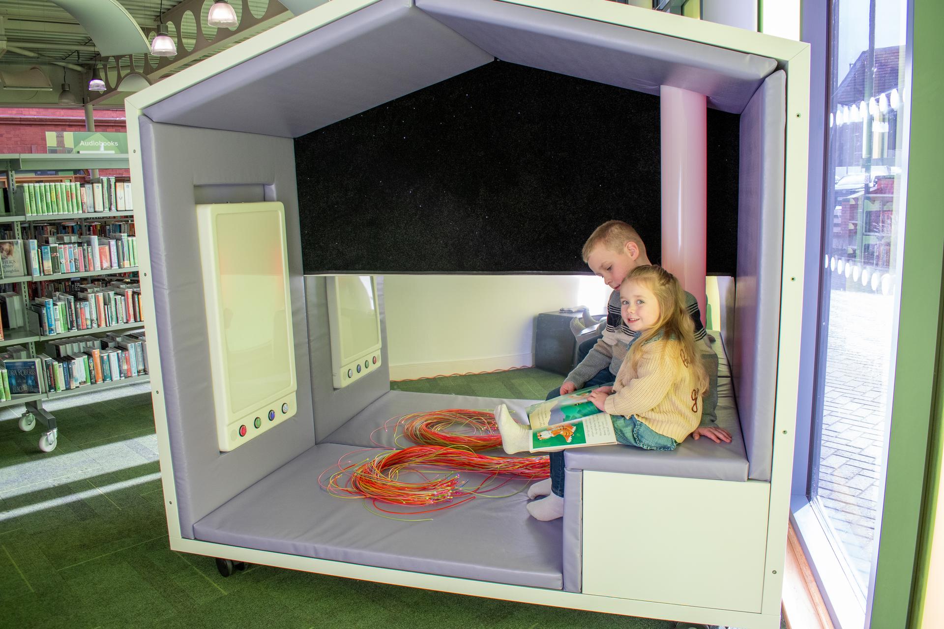 A girl and a boy are sat inside the Sensory Nook reading a book together.