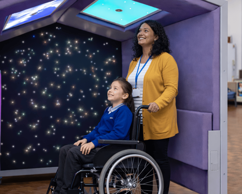 An assistant teacher and a child in a wheelchair are underneath the Sensory Shelter.