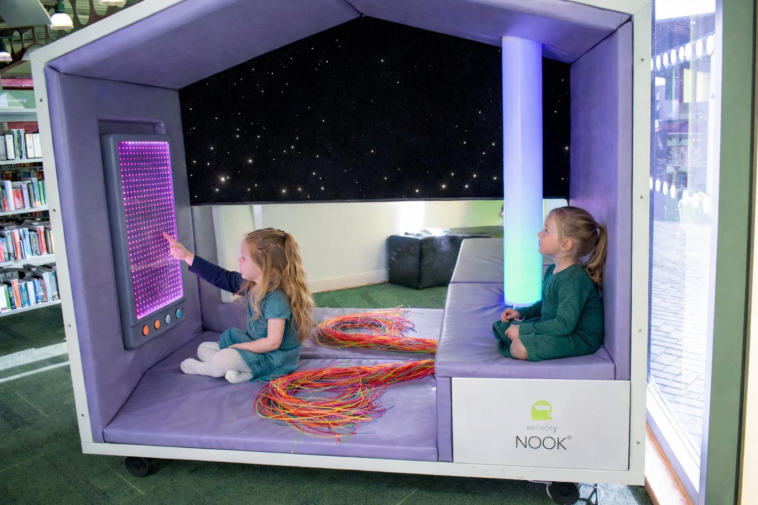 Two girls are sat inside the Sensory Nook Interactive Large, that has been placed within a Library.