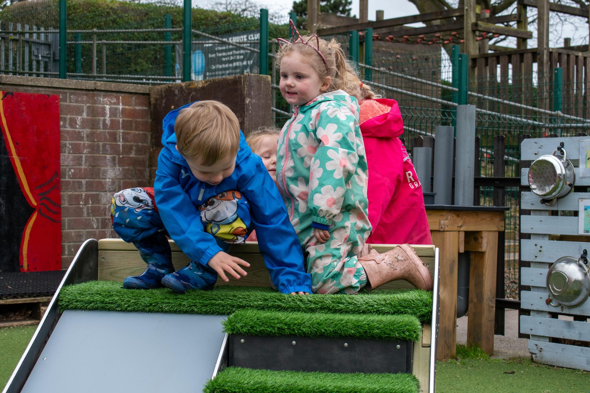 Three early years children are sat on top of the a-frame, waiting to go down the slide.