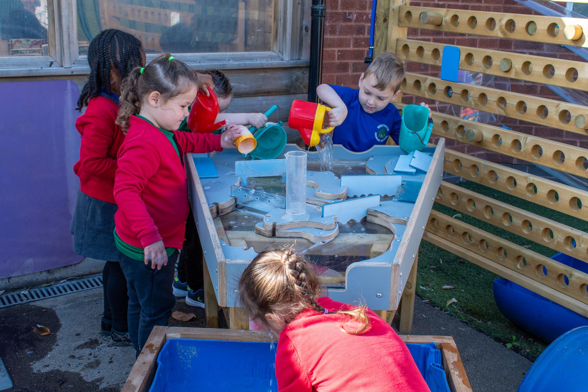 The damming station being used alongside the water wall and water table. Three children pouring water towards the top of the damming station and watching it travel to the water table