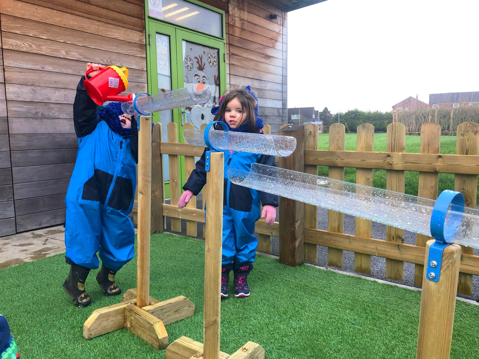 Three water channel stands with two children pouring water into them from a watering can