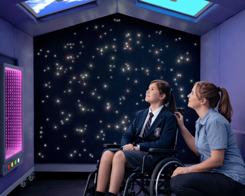 A high school girl and a teacher are both underneath the Sensory Nook Shelter, as they both look at the lighting.