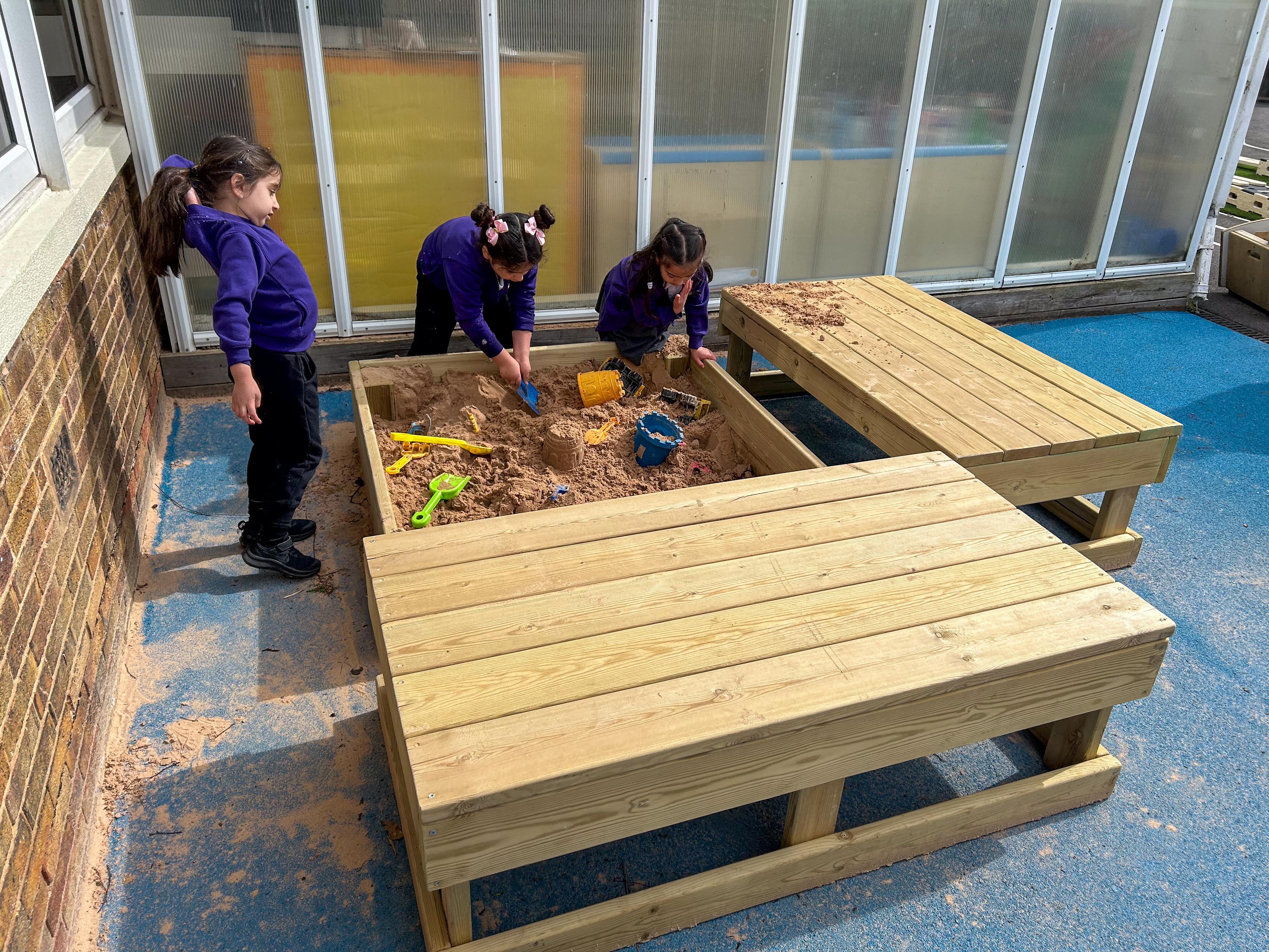 Three children are digging sand with plastic shovels in the Small Covered Sand Box.