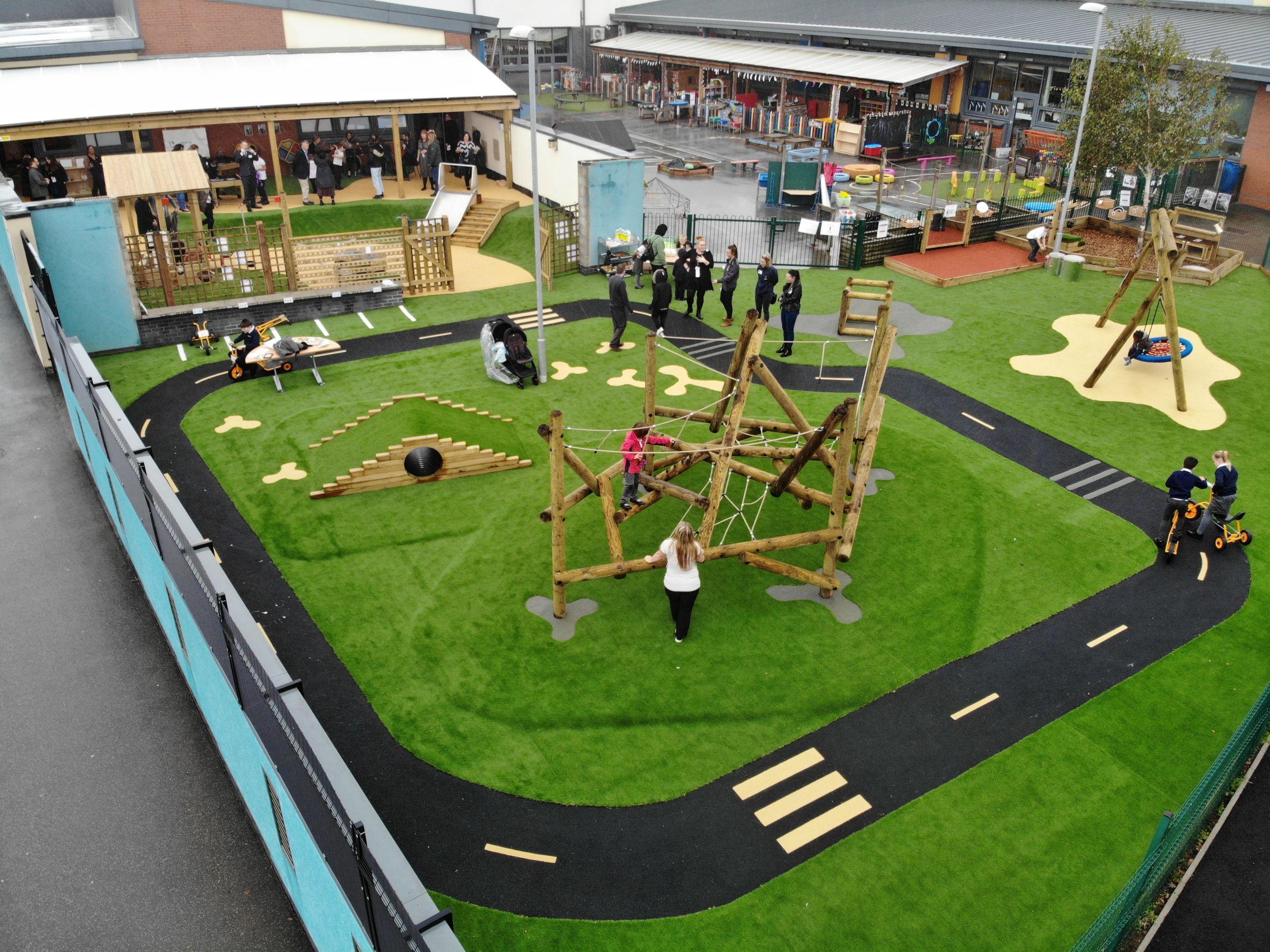 An outdoor school playground by Pentagon Play featuring a Tryfan Climber and Climb Through Tunnel hill on artificial grass and wetpour surfacing, including a black roadway track for trikes.