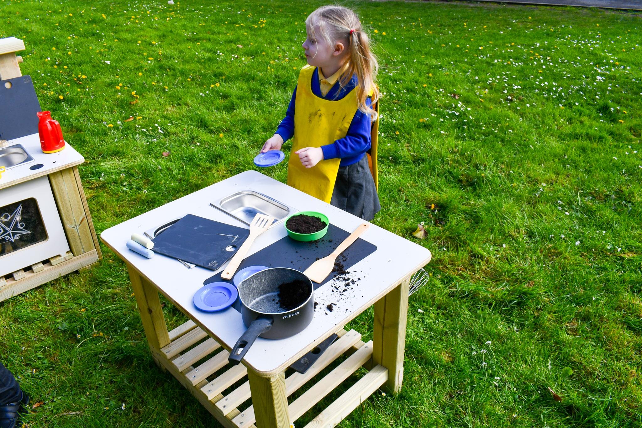 A little girl is holding a plastic bowl as she looks at the Mud Kitchen Island.