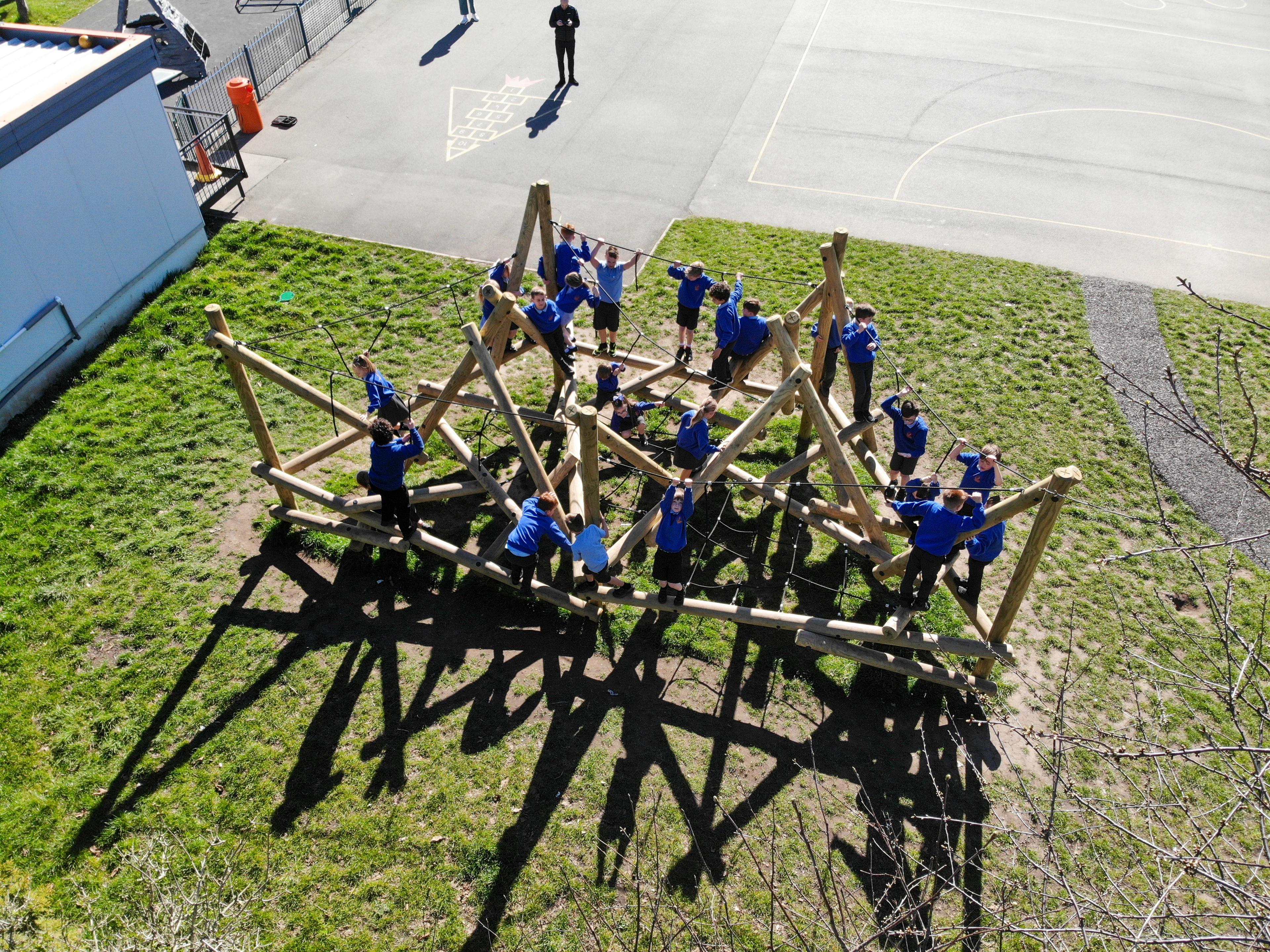 A group of school children in blue jumpers playing on a large wooden climbing frame in a playground.