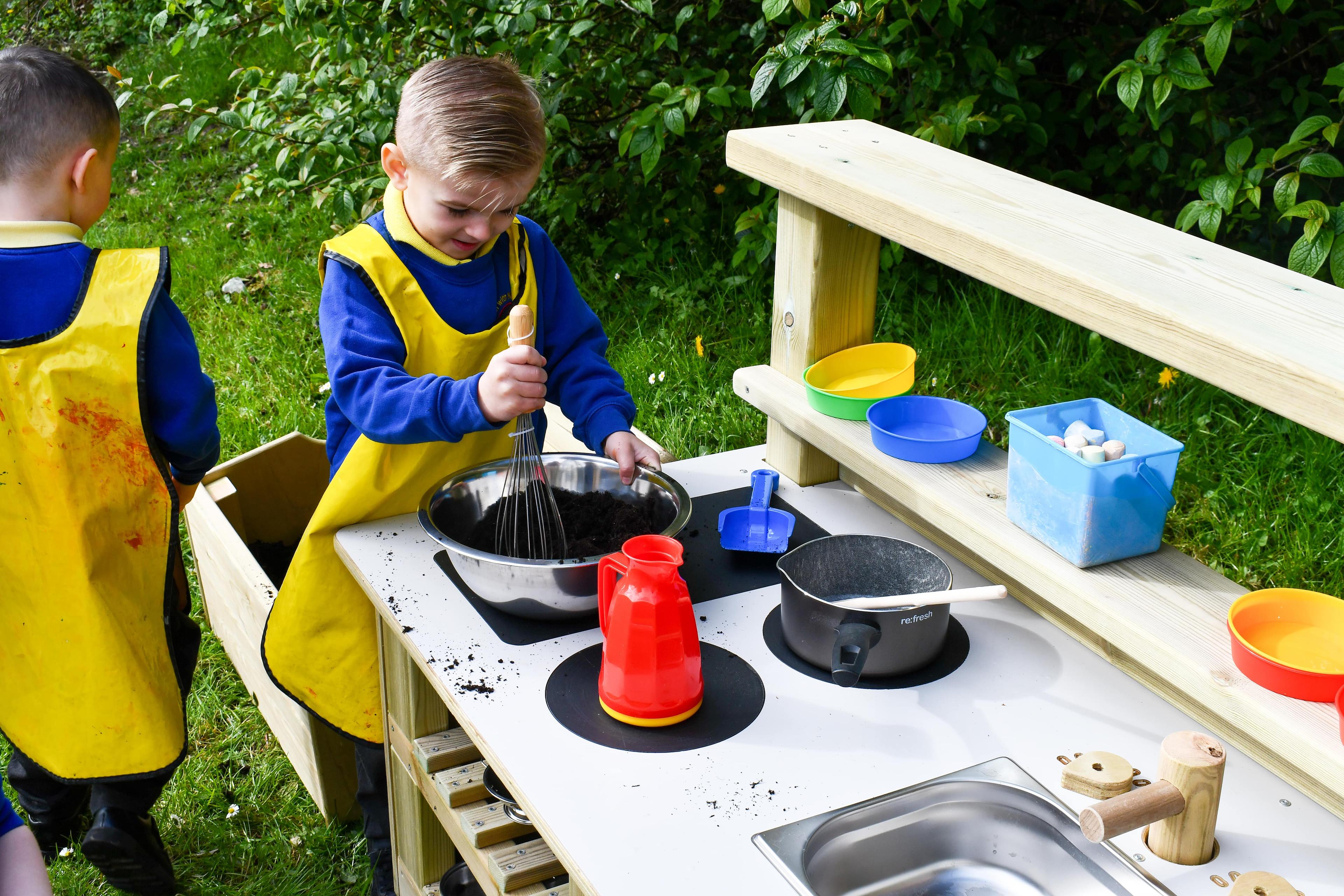 A boy is mixing mud in a bowl that has been placed on the Wooden Mud Kitchen.