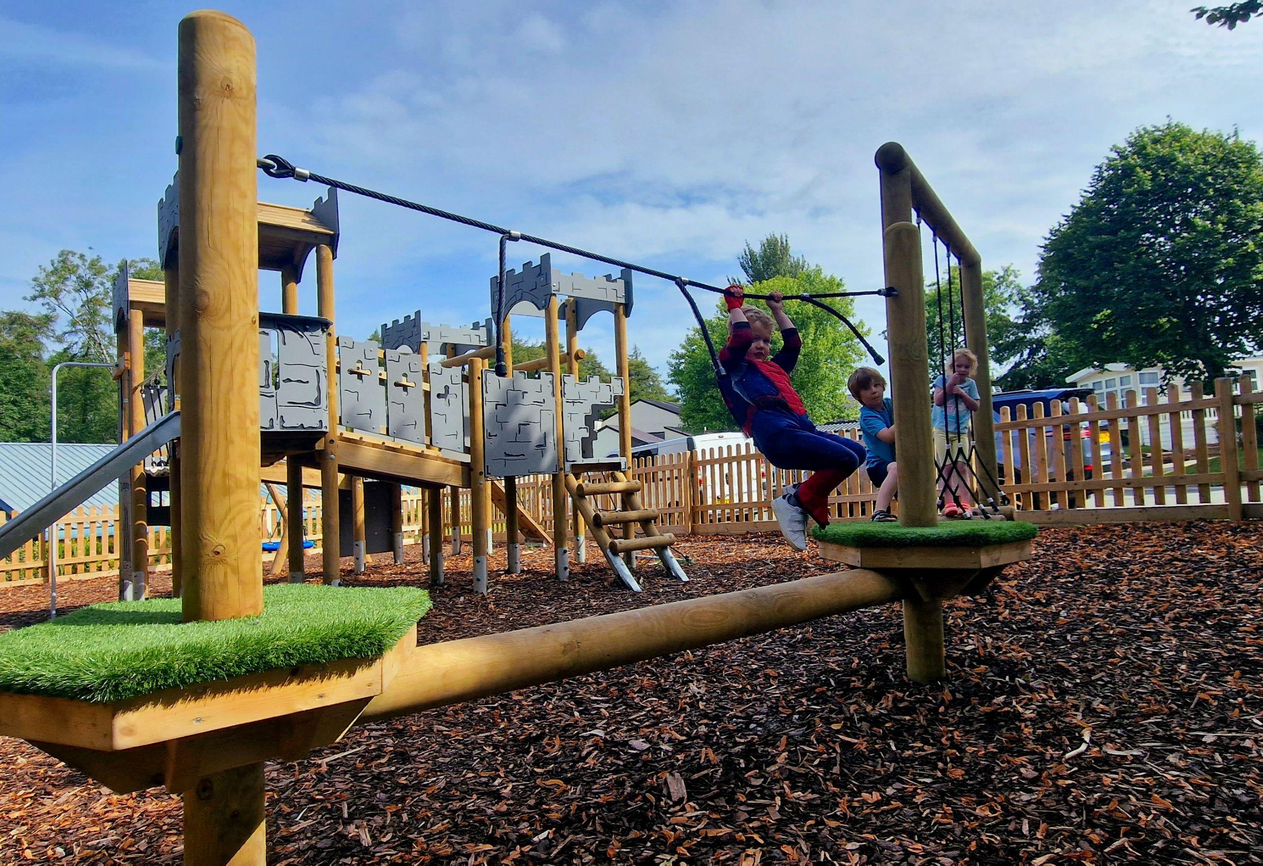 A child swings on a platform to platform beam crossing, part of a trim trail at Brynteg Holiday Park, with a bespoke castle-themed play tower in the background on wood chip surfacing.