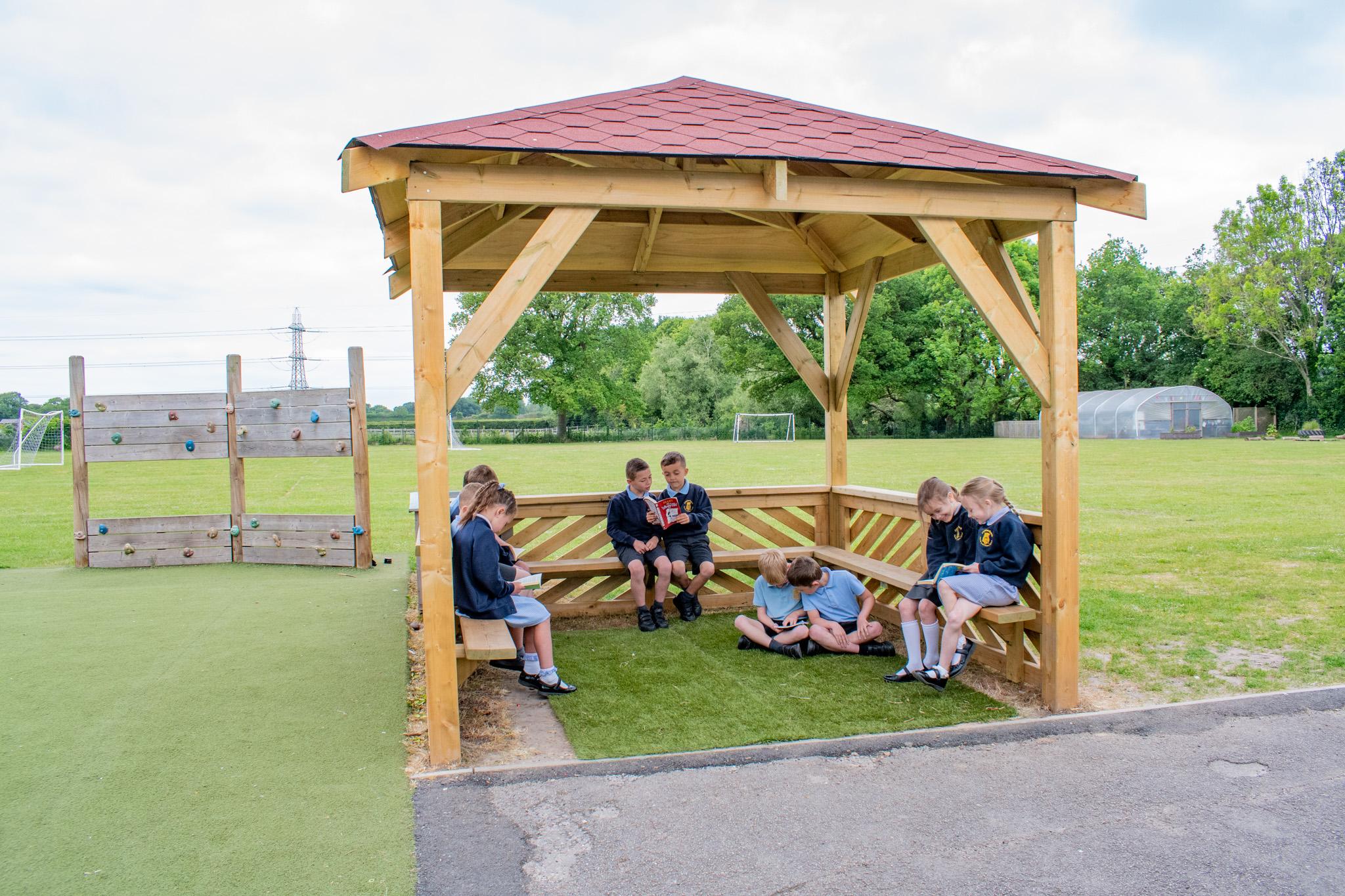 A group of 8 school children in blue uniform, sitting on wooden benches within a in wooden 3m gazebo, pitched on grass, outside a school playing field.