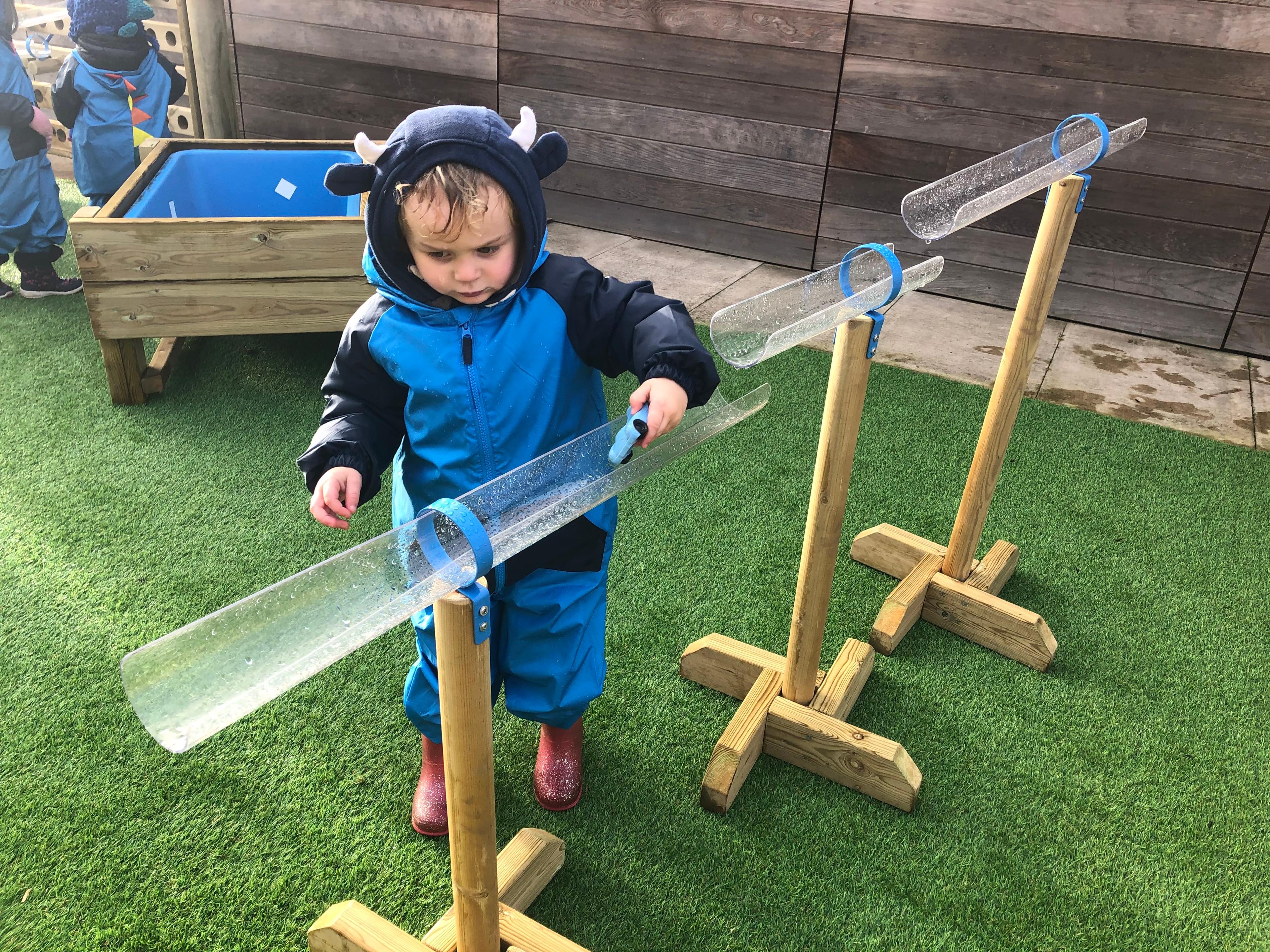 A little boy is driving a toy car through the plastic tubes that are held up by the water play stands.
