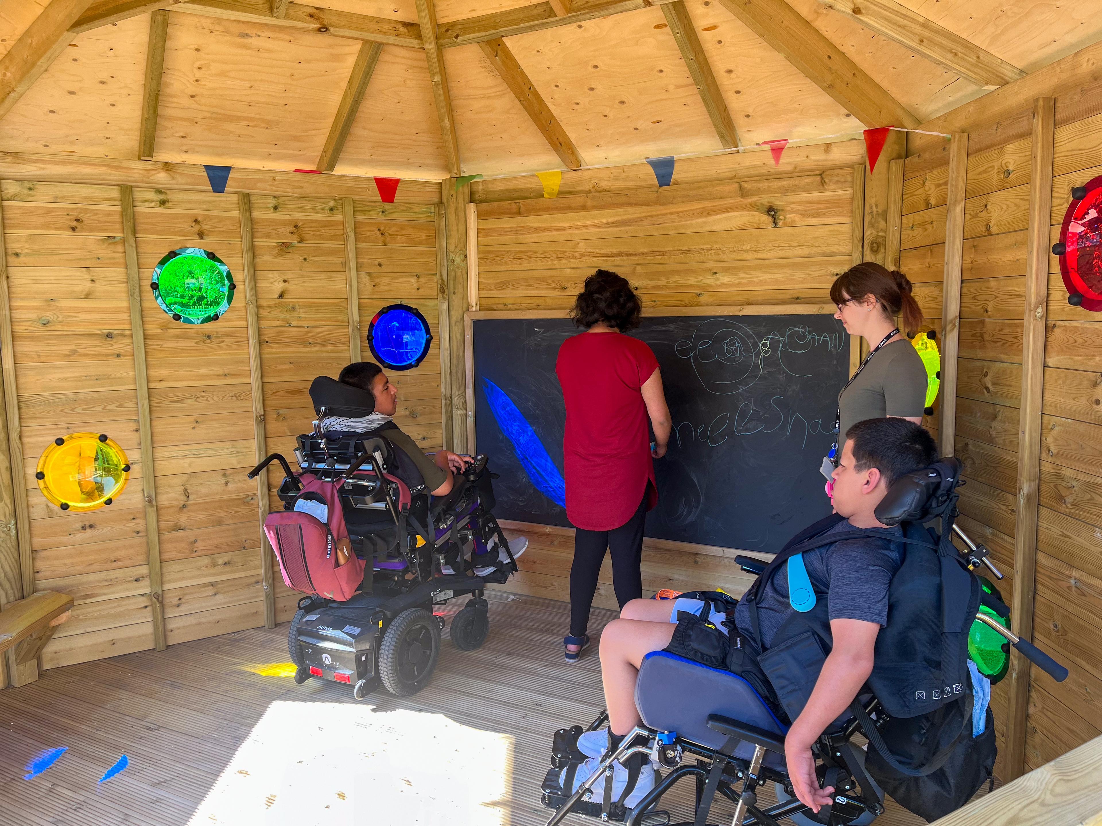 Children in wheelchairs with school staff writing on the whiteboard inside the Sensory 5M Hexagonal Gazebo