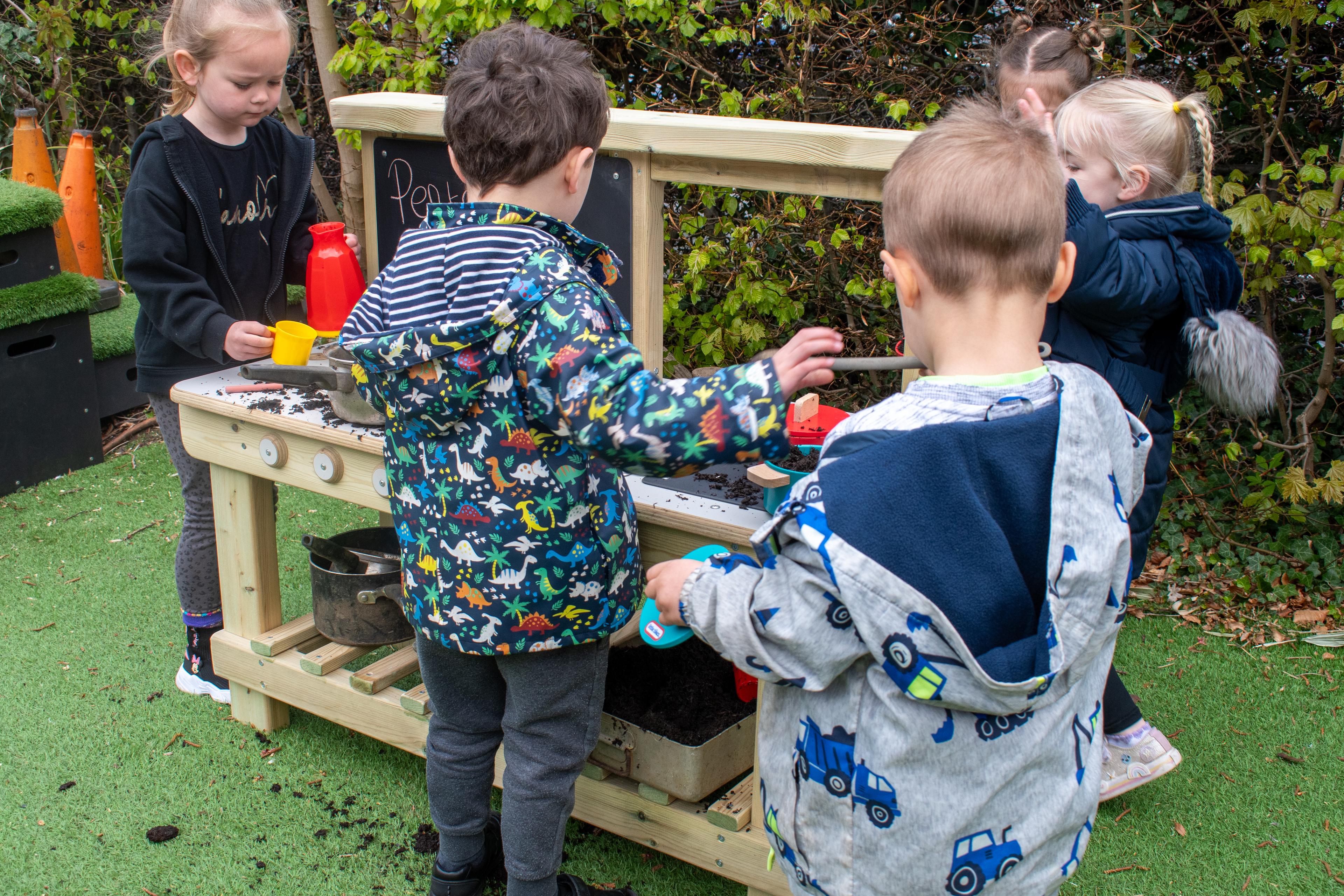 A group of EYFS children are surrounding a mud kitchen and playing with it.