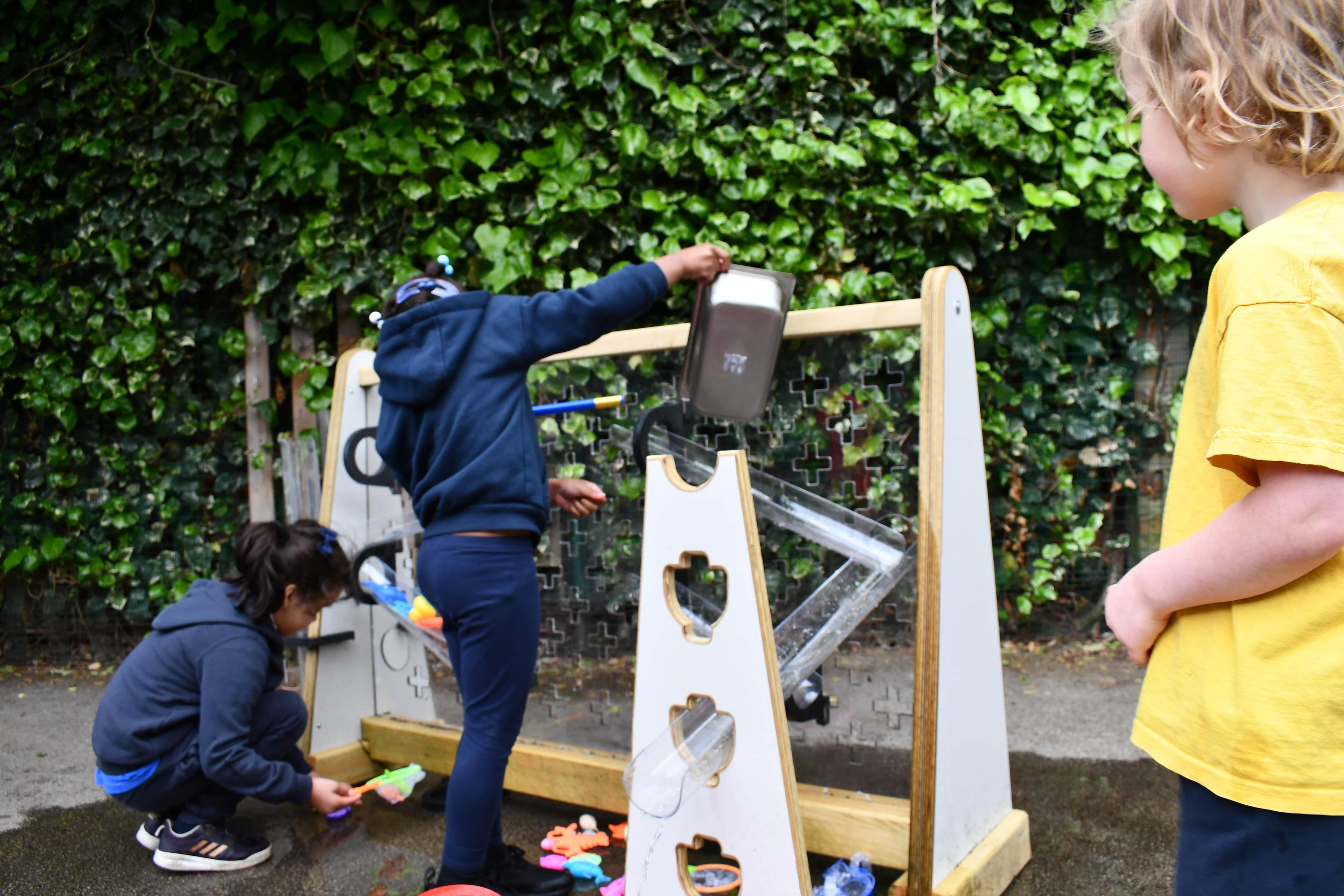 A small group of children are pouting water down the freestanding water wall, as the pipes go through a channel stand.