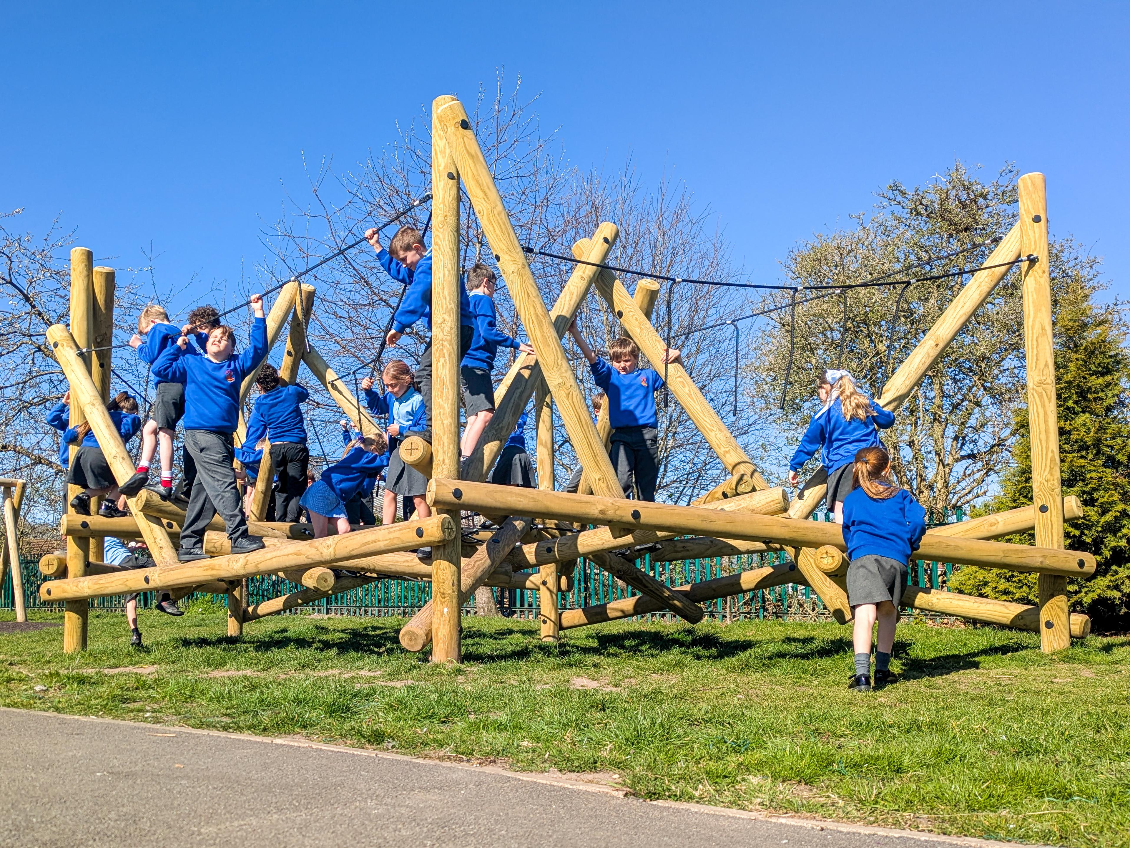 A group of school children in blue jumpers playing on a large wooden climbing frame in a playground.