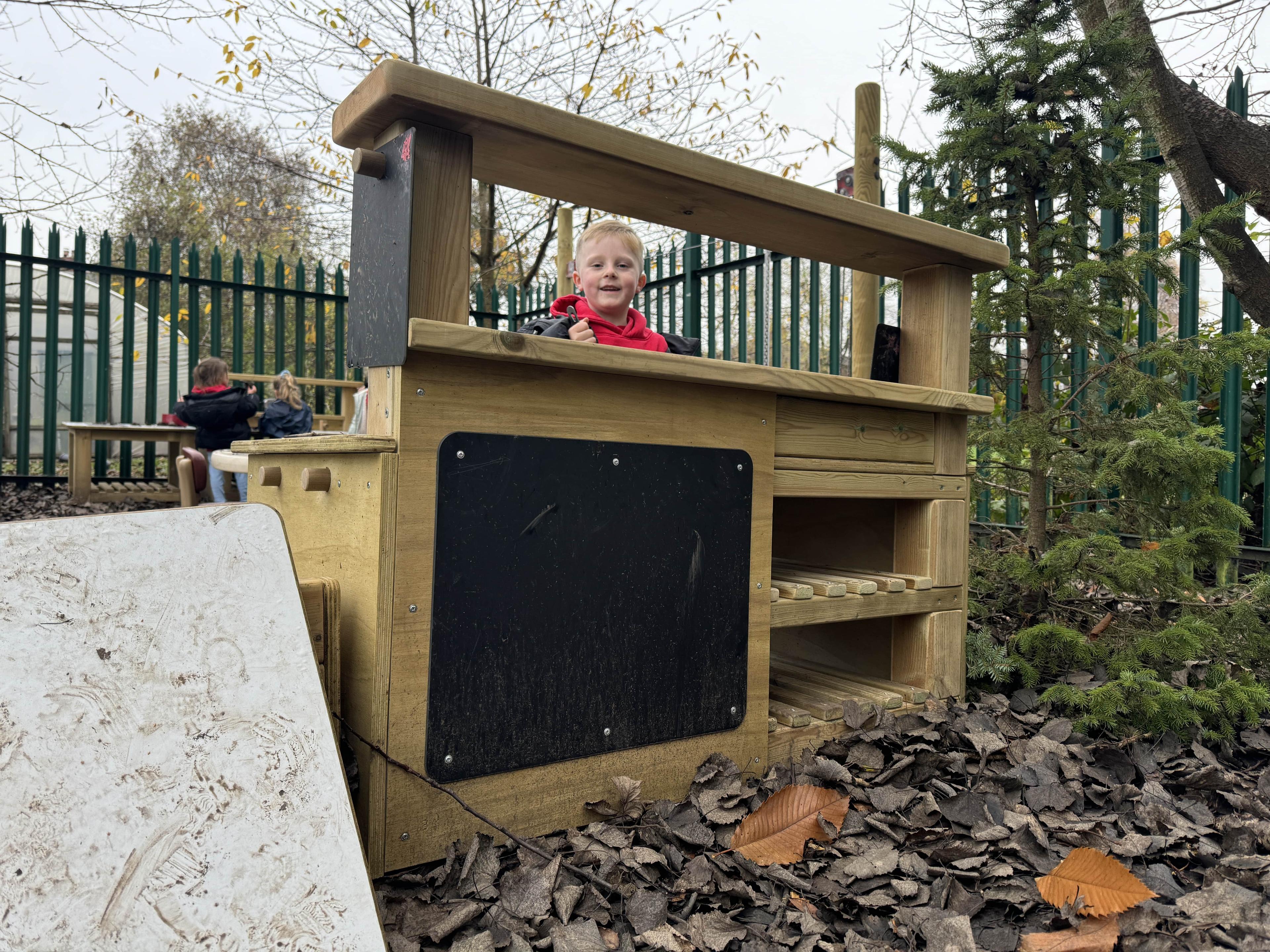A boy is nursery is looking over the Wooden Mud Kitchen towards the camera.