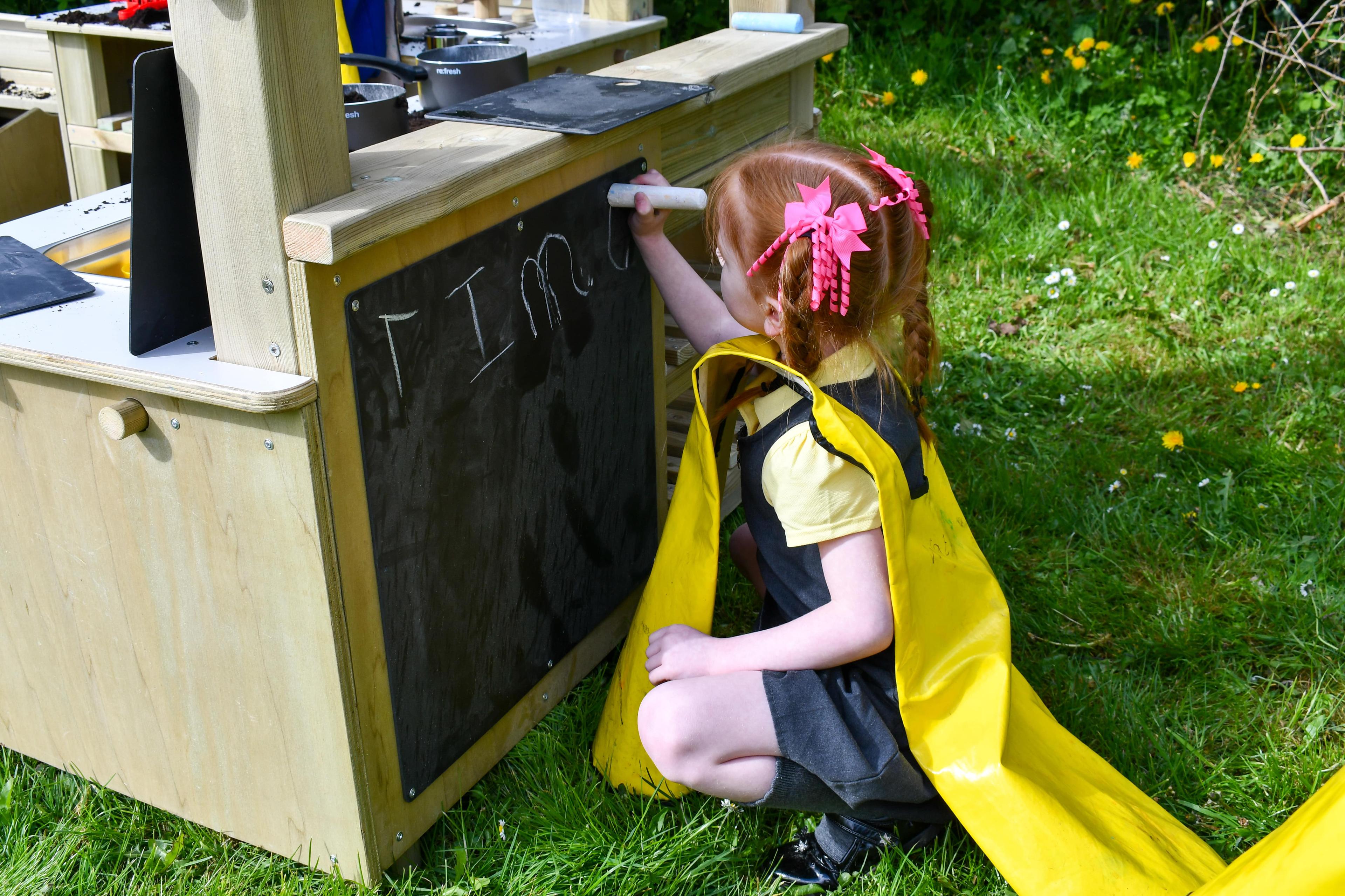 A little girl is writing something on the chalkboard that has been placed on the back of the Wooden Mud Kitchen.