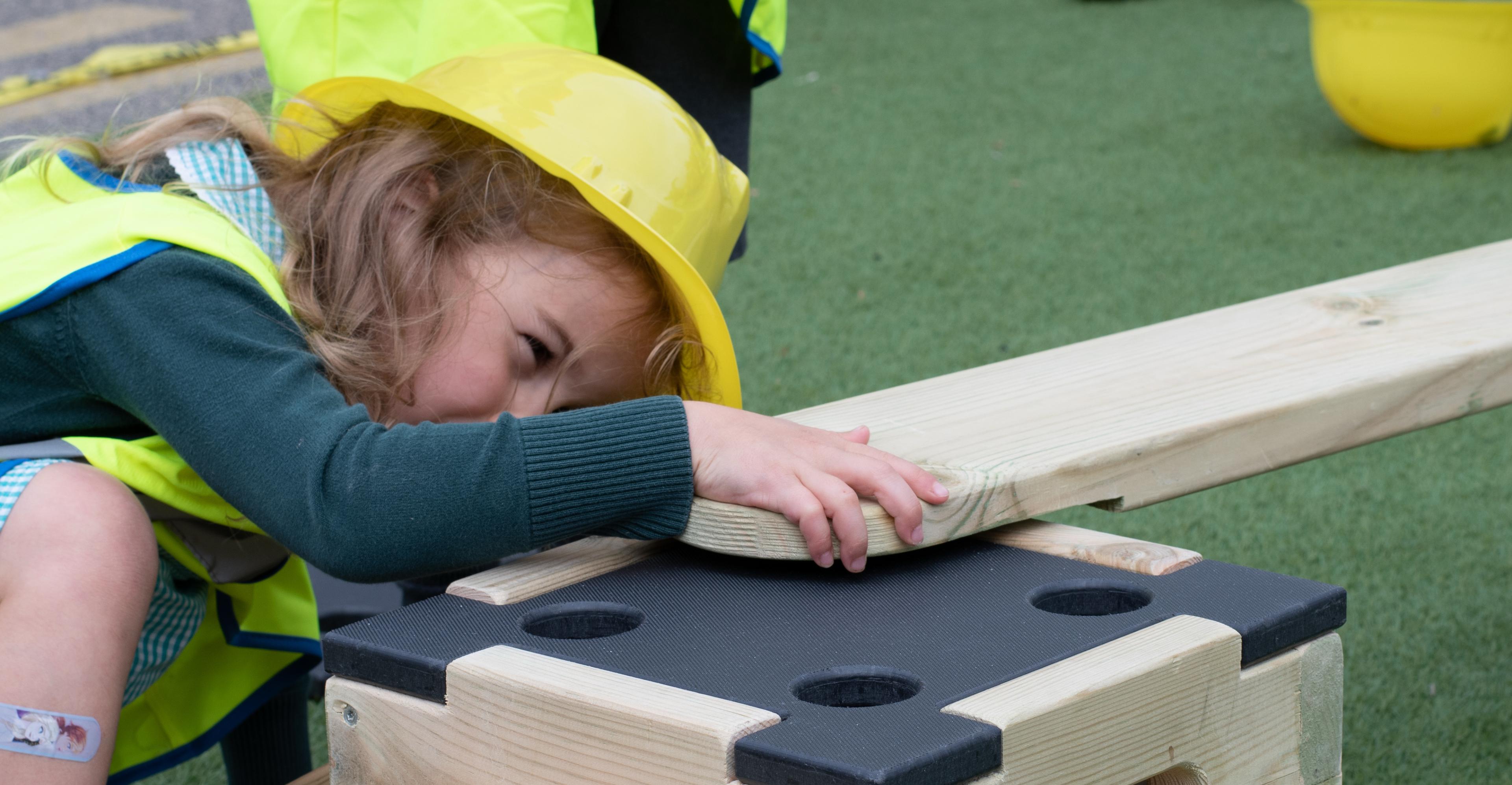 A young student wearing a hardhat and hi-vis jacket attaching 2 sections of Pentagon Play's Play Builder together