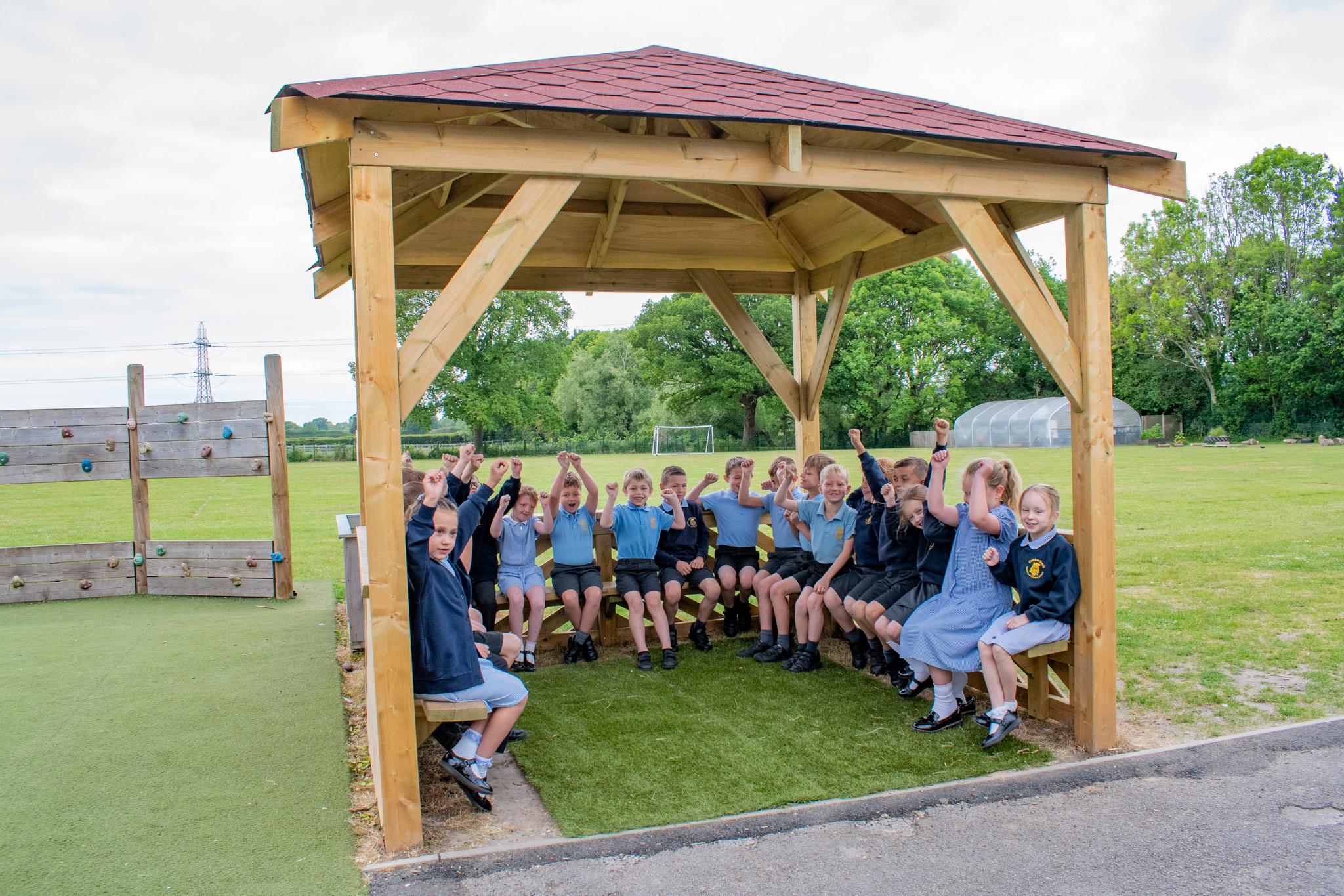 A group of 15 school children in blue uniform, sitting on wooden benches within a in wooden 3m gazebo, pitched on grass, outside a school playing field.