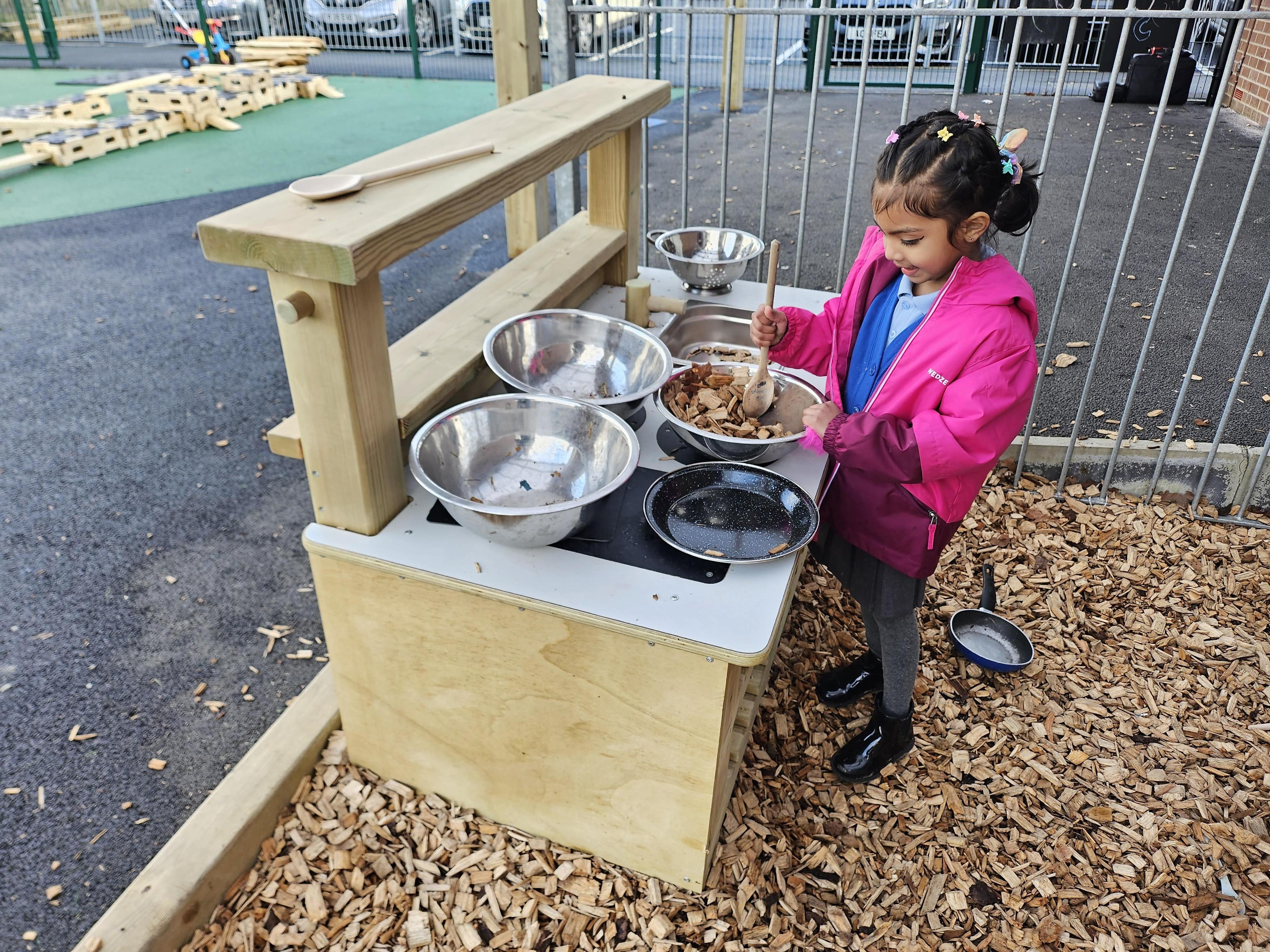 A girl in nursery is mixing mud in a metal bow on the surface of the Wooden Mud Kitchen.
