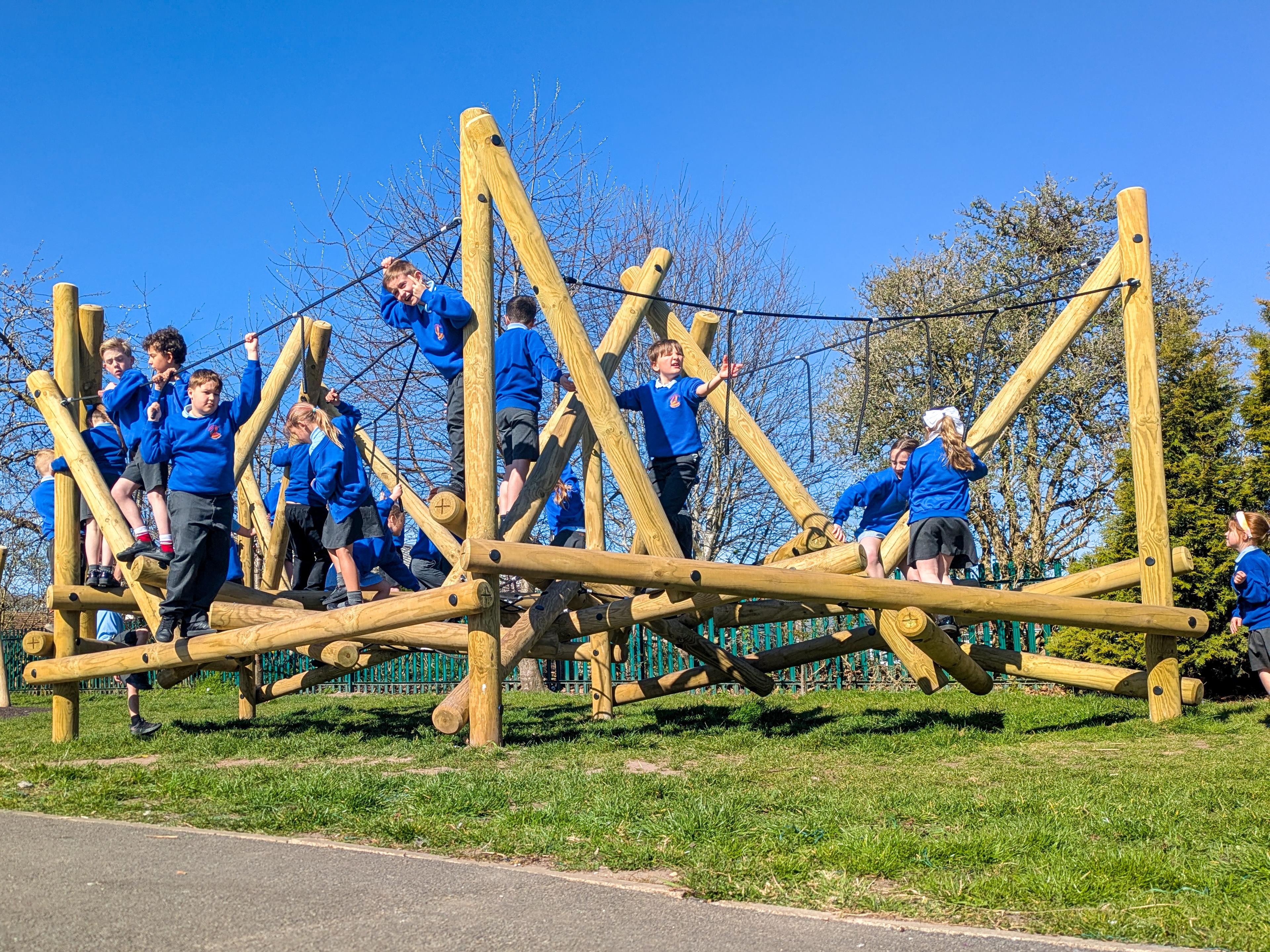 A group of school children in blue jumpers playing on a large wooden climbing frame in a playground.