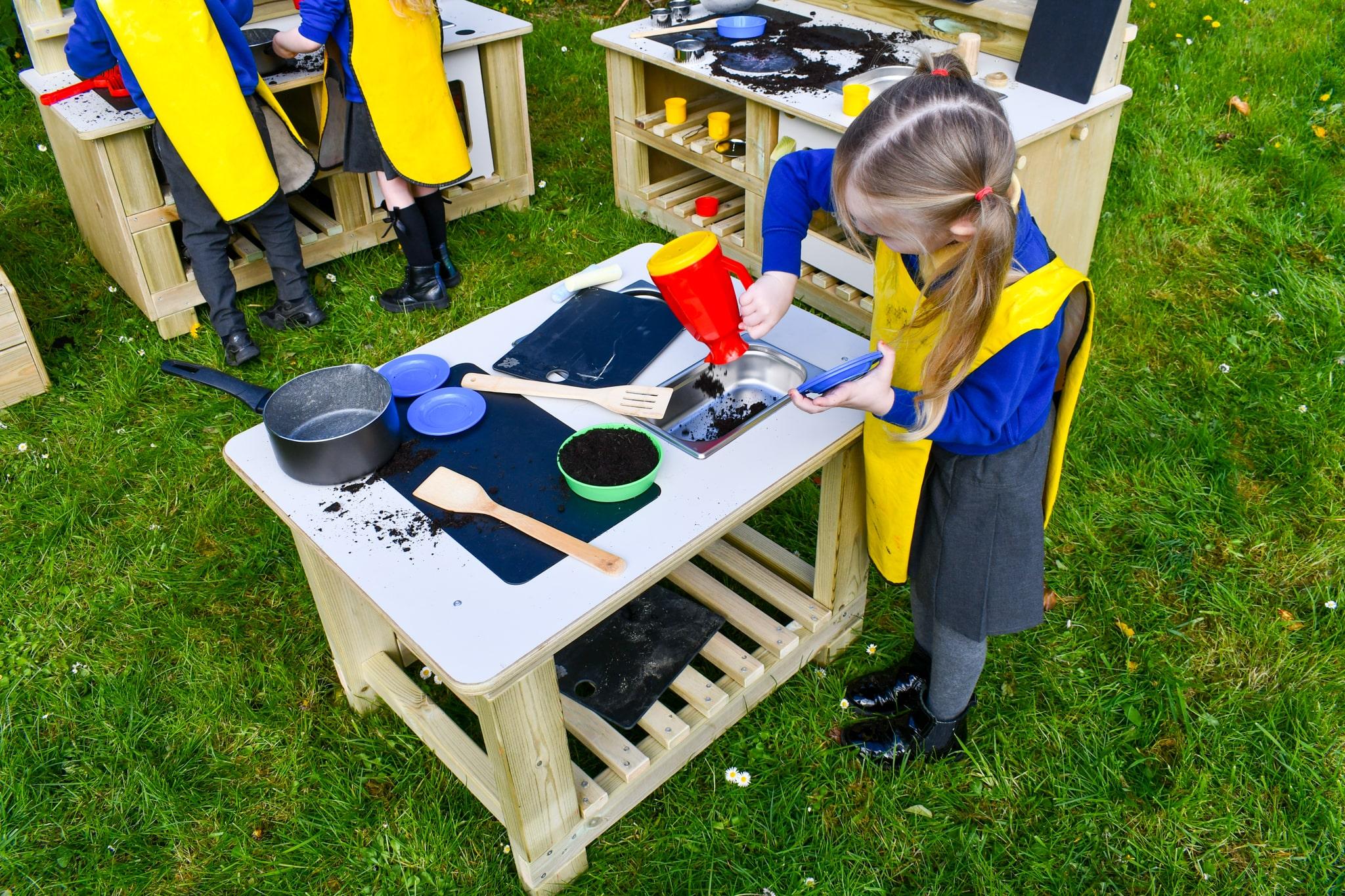 A little girl is pouring mud from a toy kettle into the Mud Kitchen Island's sink.