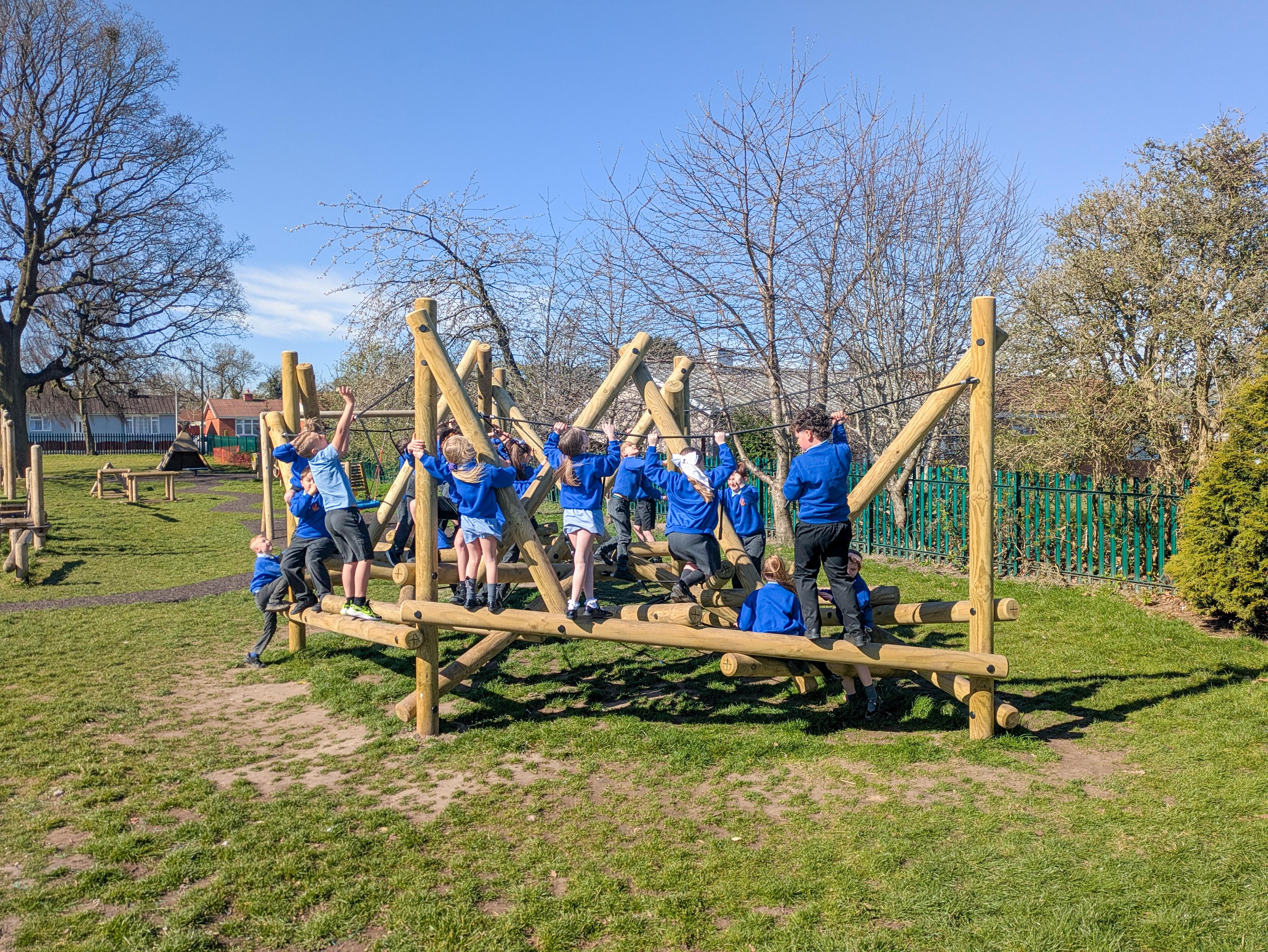 A group of school children in blue jumpers playing on a large wooden climbing frame in a playground.