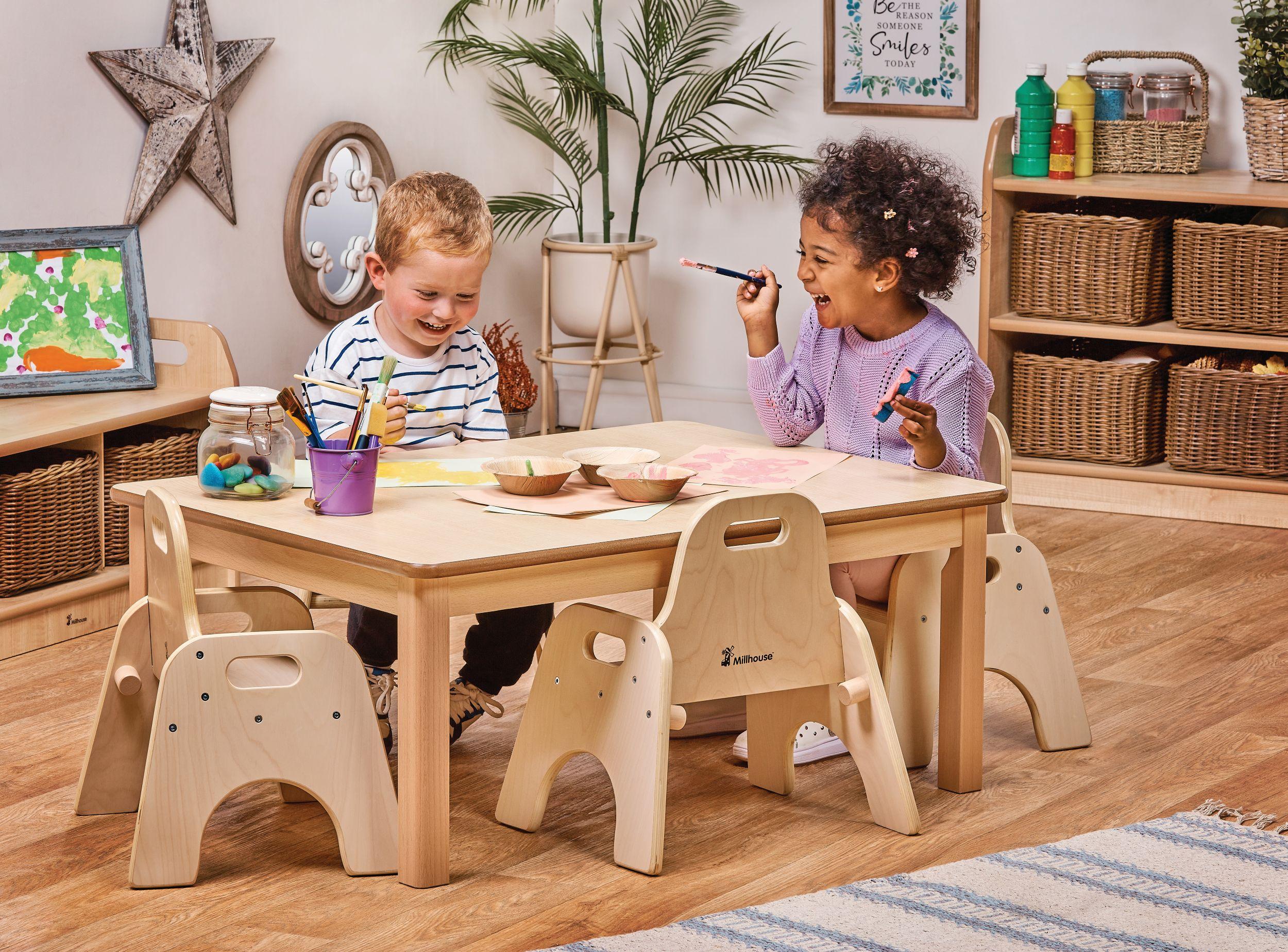 Two children laugh while painting at a Millhouse natural timber table with matching ergonomic chairs in a bright, modern classroom filled with organized craft supplies and greenery.