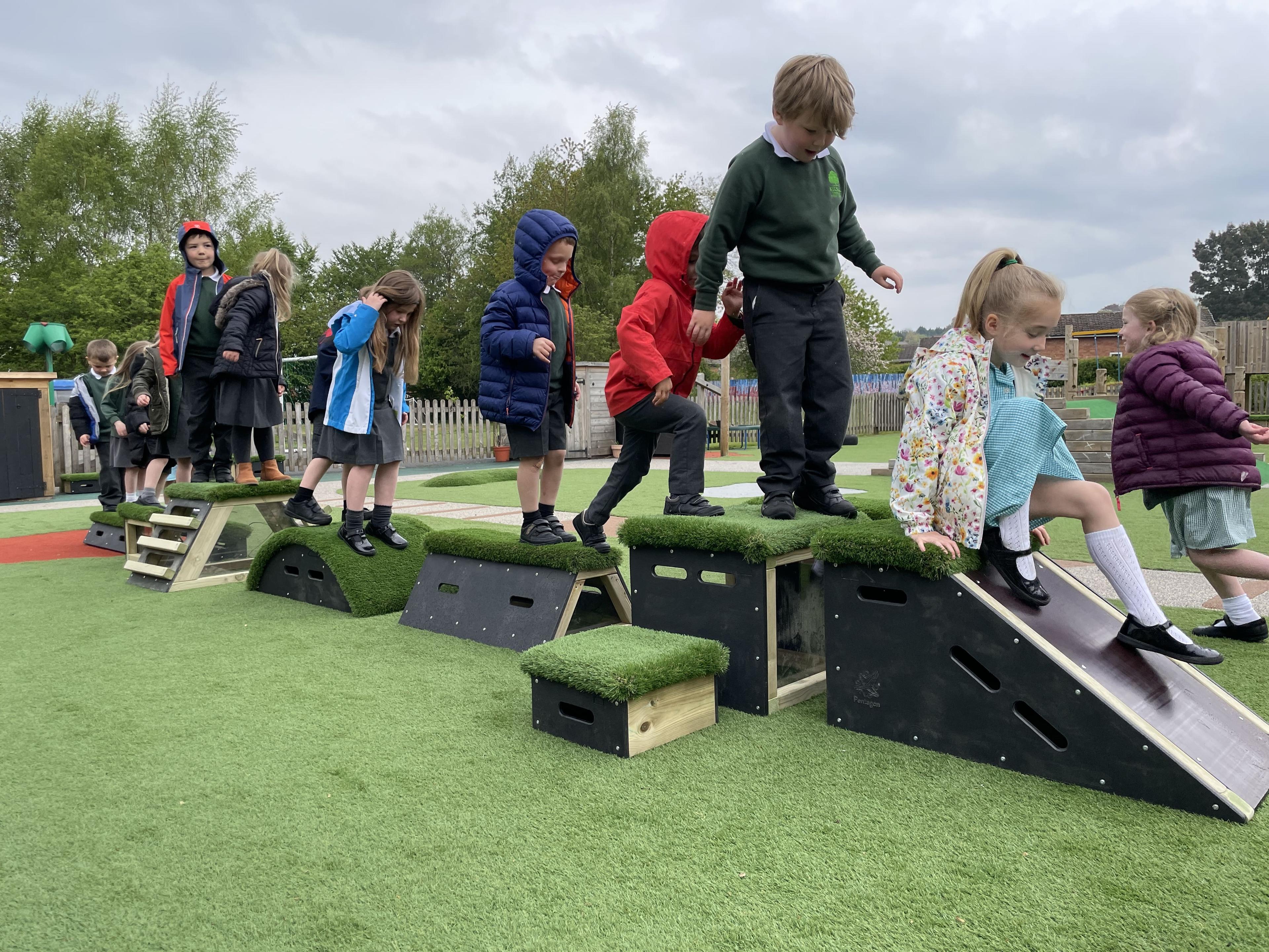 A line of reception children are stood on the different blocks including within the Peaks Set, as they wait behind a girl sliding down the Slide block.