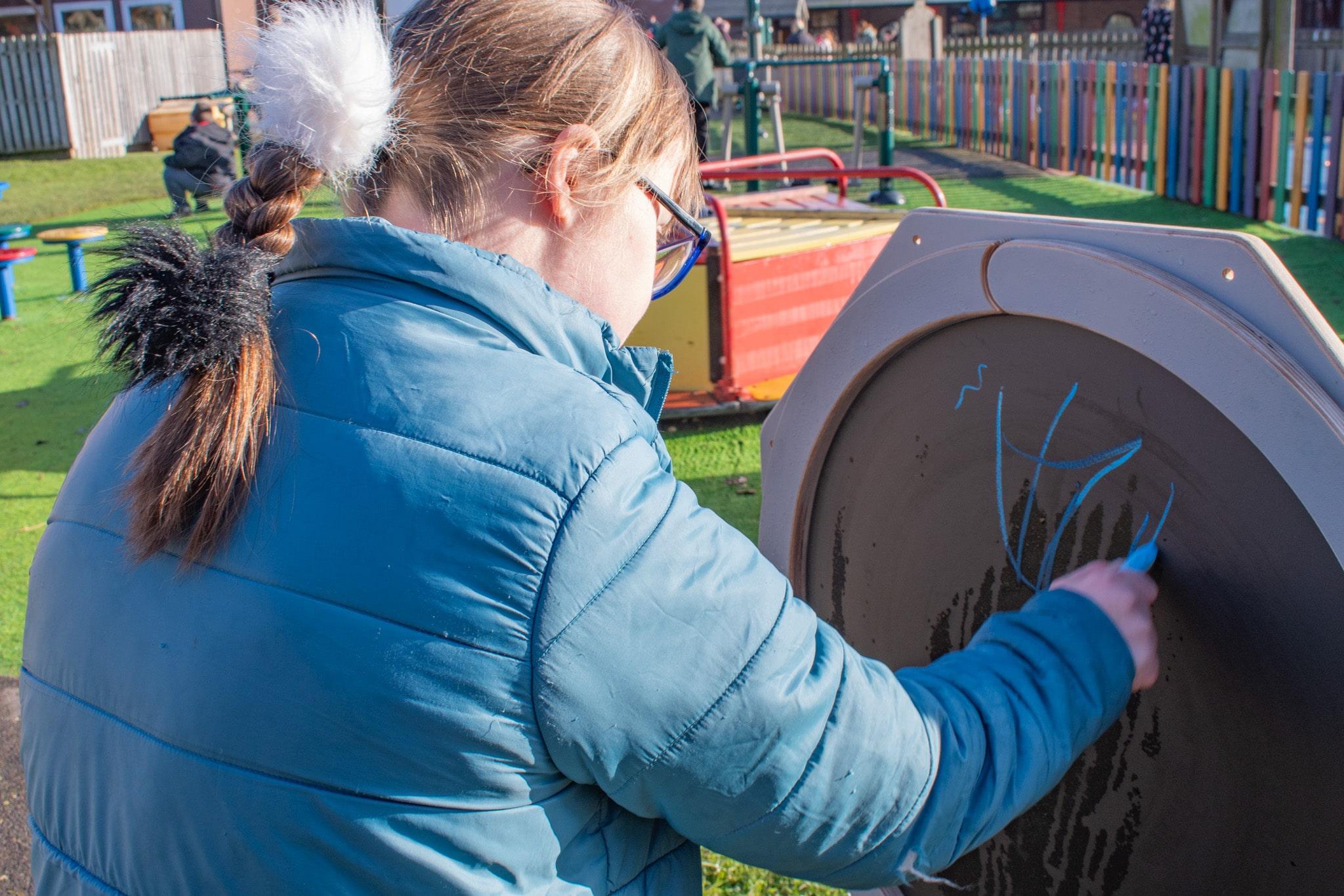 A girl is drawing on the chalkboard attached to the Drawing Sensory Spinner with blue chalk.