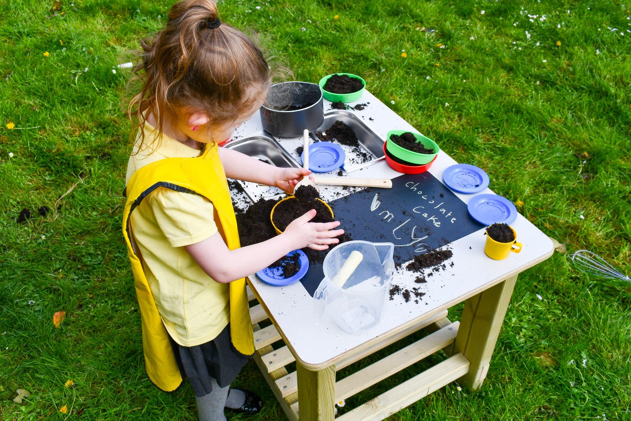 A little girl is patting mud into a bowl on the surface of the Mud Kitchen Island.