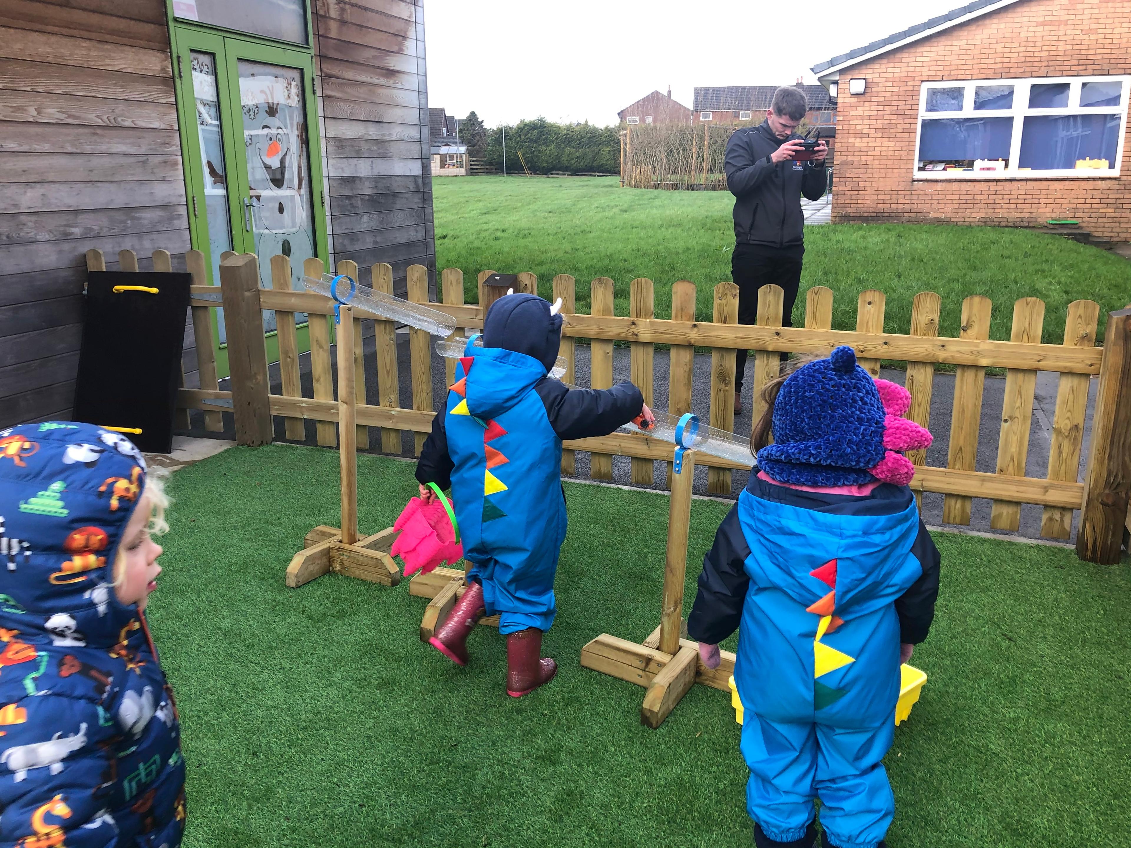 Three children are playing with the Water Channel Stand Set as an adult can be seen controlling a drone.