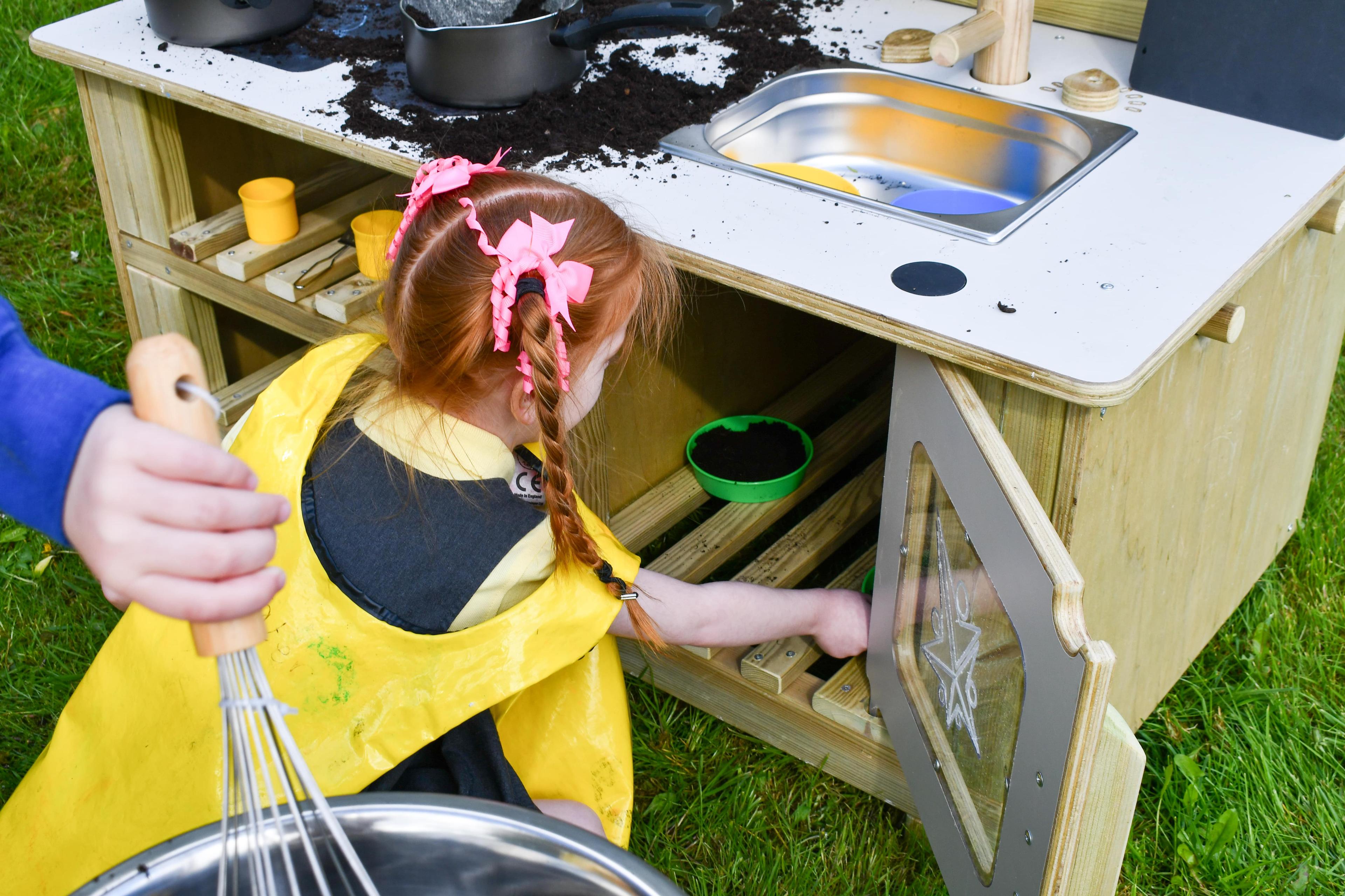 A little girl is putting mud pies into the oven on the Wooden Mud Kitchen.