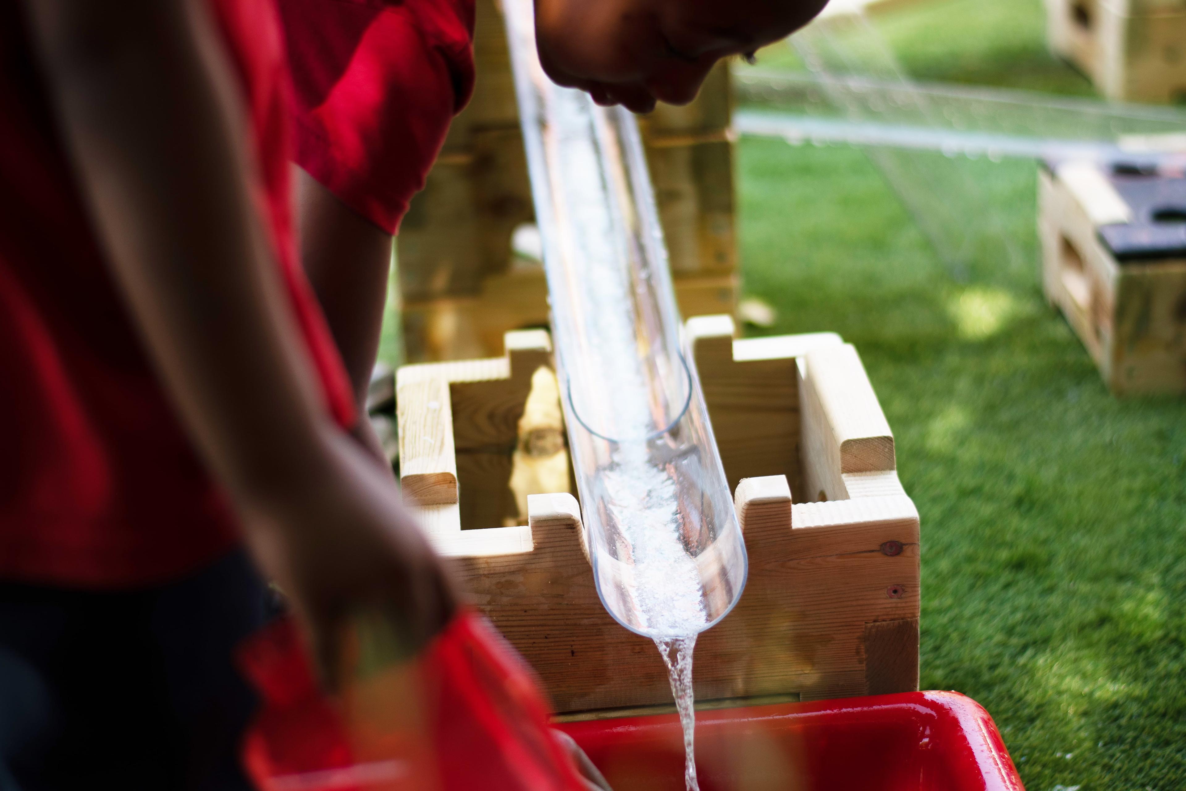 A girl is collecting water from the plastic pipes of the Play Builder Water Play set in a red plastic container.