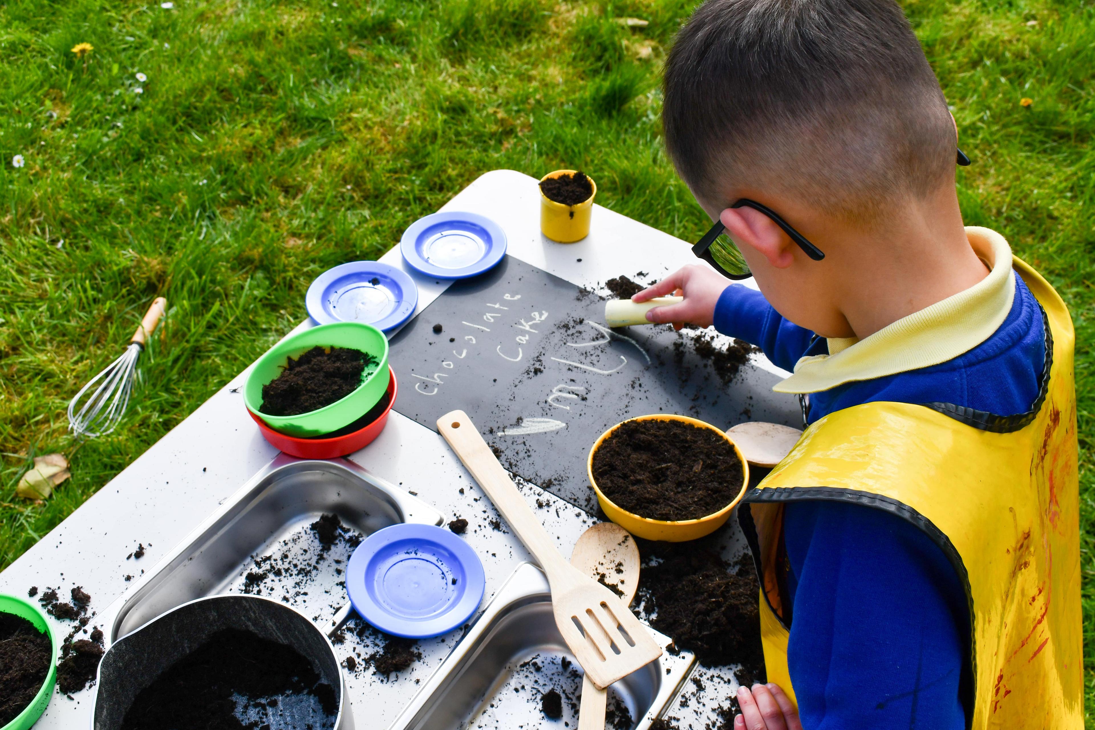 A little boy is writing a recipe on the chalkboard attached to the surface of the Mud Kitchen Island.