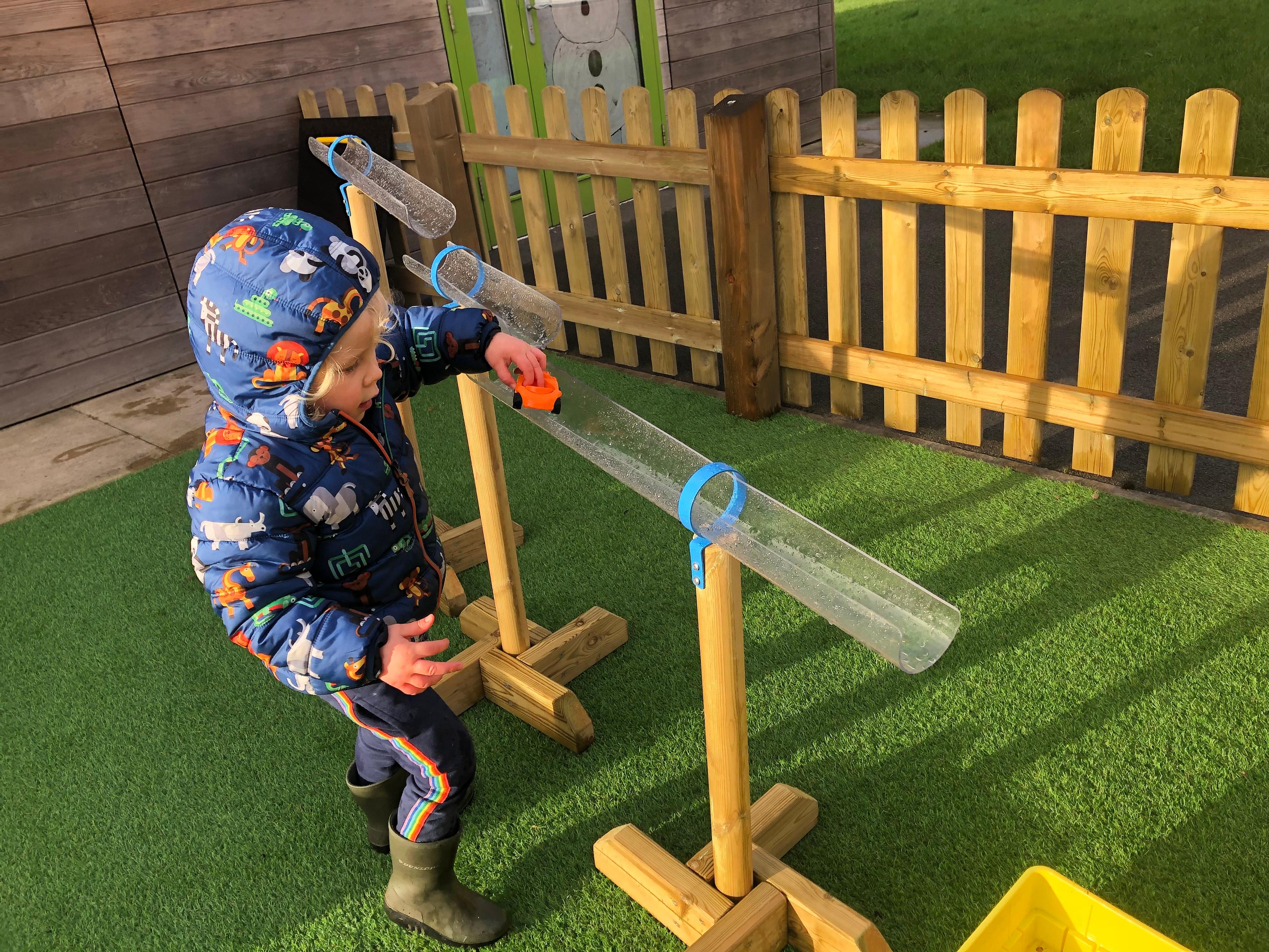 A EYFS boy is driving a toy car through the plastic tubes that are held up by the water play stands.