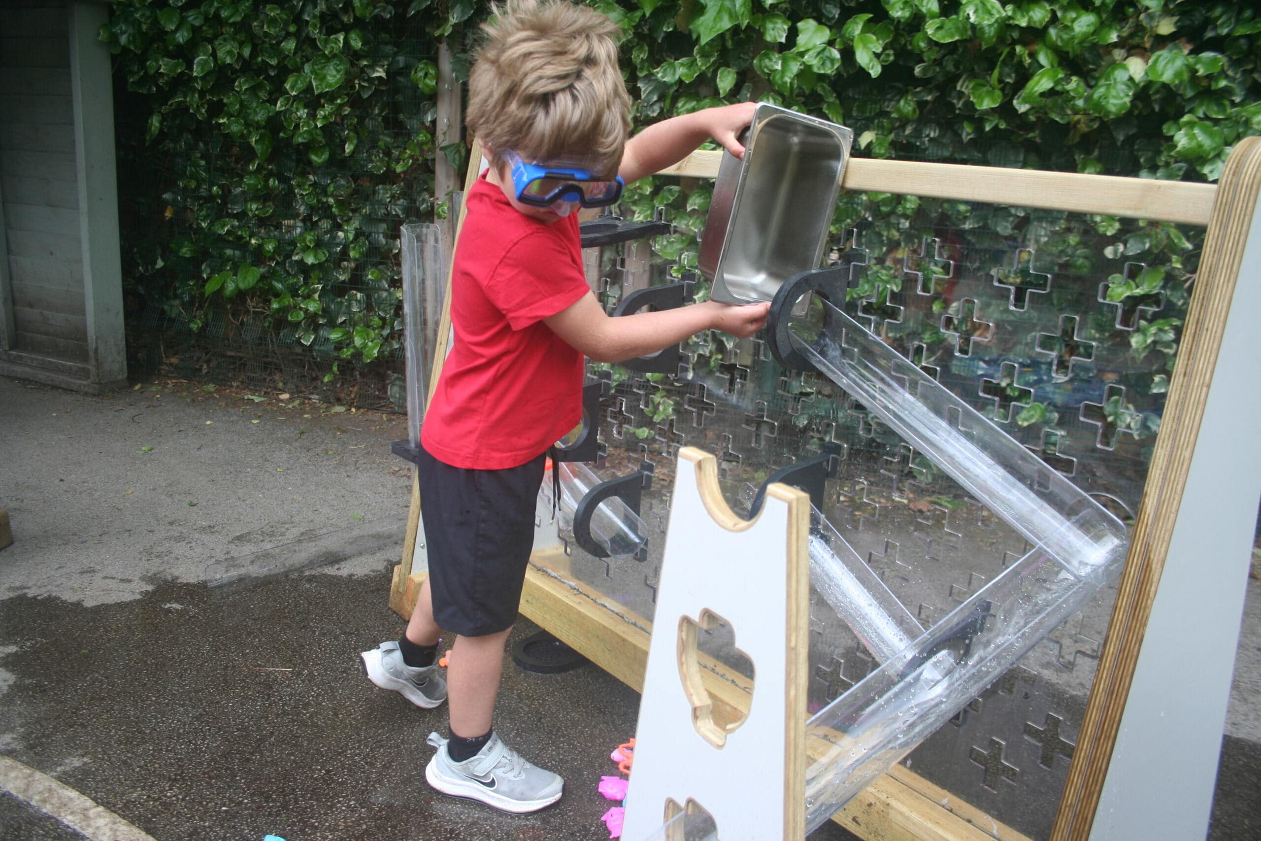 A little boy wearing goggles is pouring water down the water wall as he watches it flow.