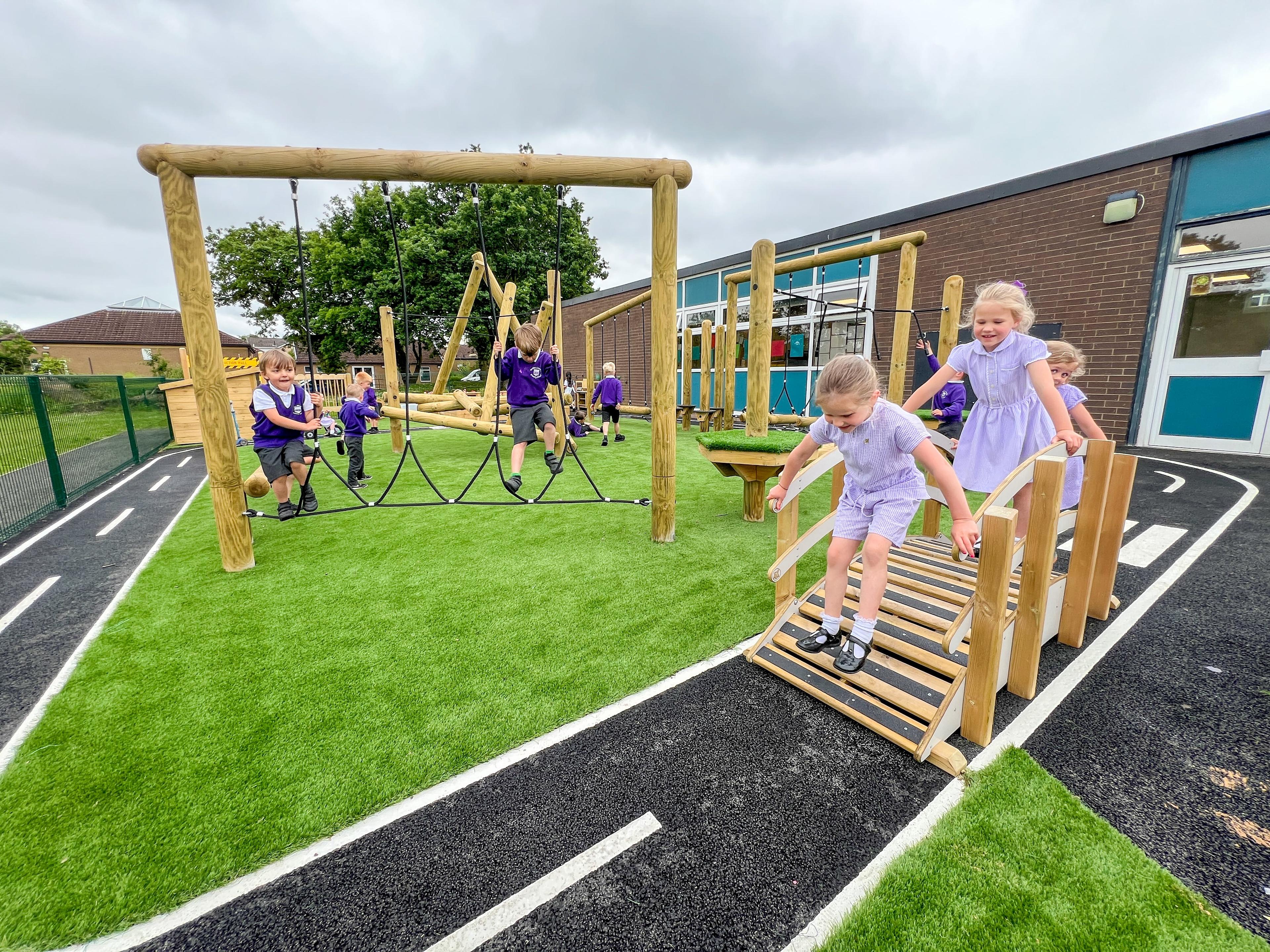 A small group of early years children are crossing the Play Bridge with Handrails as another group of children are playing on a trim trail.