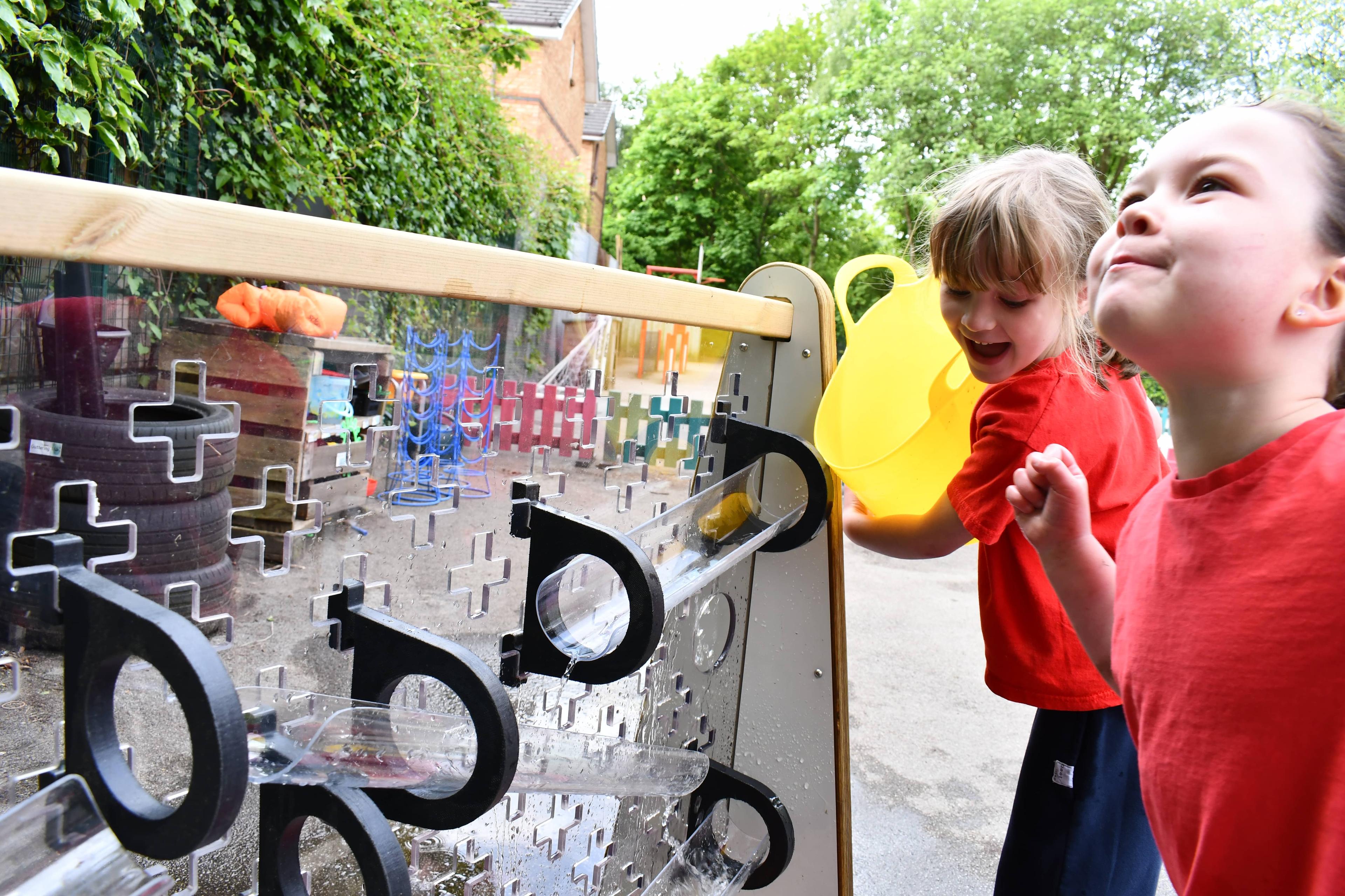 Two little girls are watching the freestanding early years water wall as water flows down it.