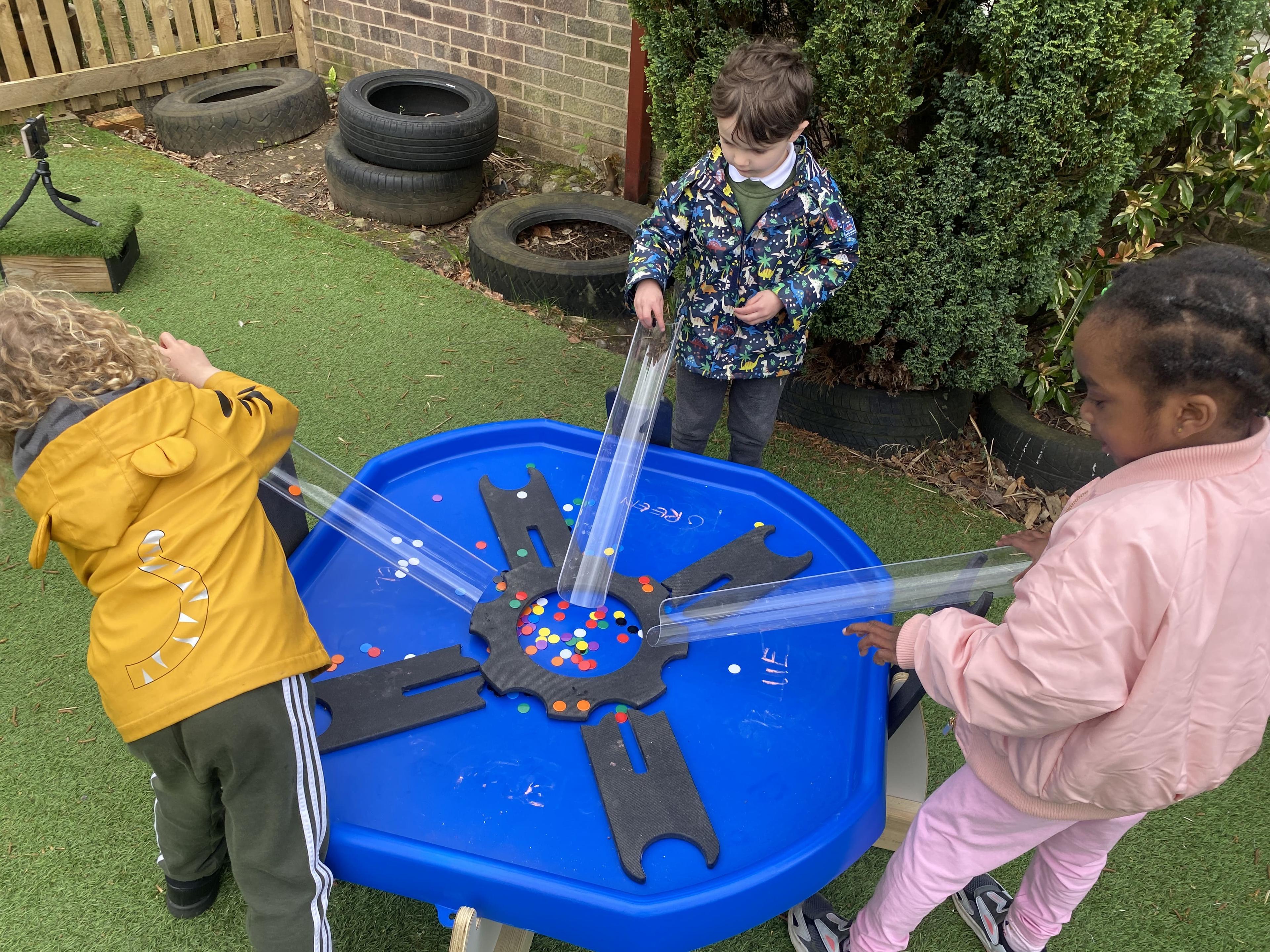 Three children are putting counters down plastic tubes that are connected to a tuff spot table.