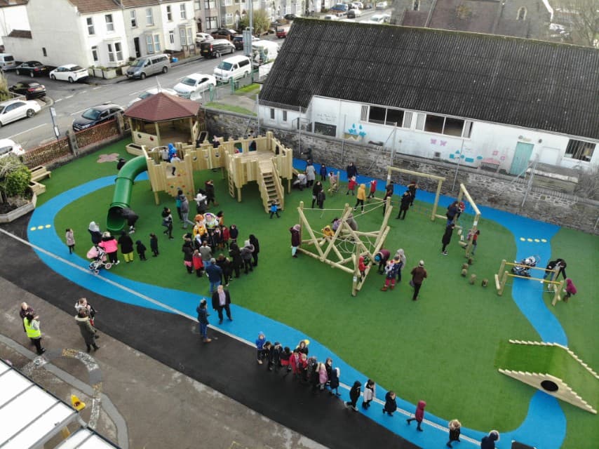 A birds-eye shot of a playground with a variety of active play equipment. There's a Play Tower, a climbing frame and a ramp. The equipment is installed on top of artificial grass with a blue wetpour river design going around the area.