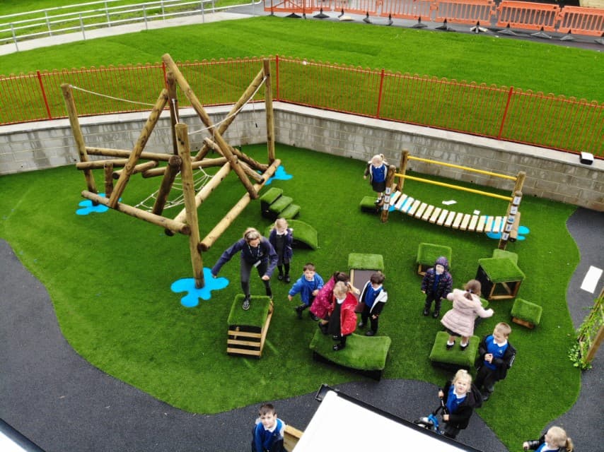 A play area shown from a birds-eye view. A wooden climbing frame, a set of Get Set, Go blocks and a small wooden bridge are placed on top of an artificial grass surface. A group of children can be seen playing with all the pieces of equipment.