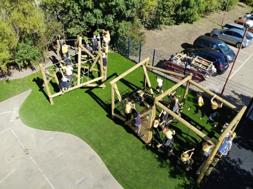 A play area from a birds-eye view. Two pieces of play equipment are installed on top of the surface, one being a climbing frame and the other being a Forest Trim Trail. A group of children can be seen playing on both of these pieces of active play equipment.