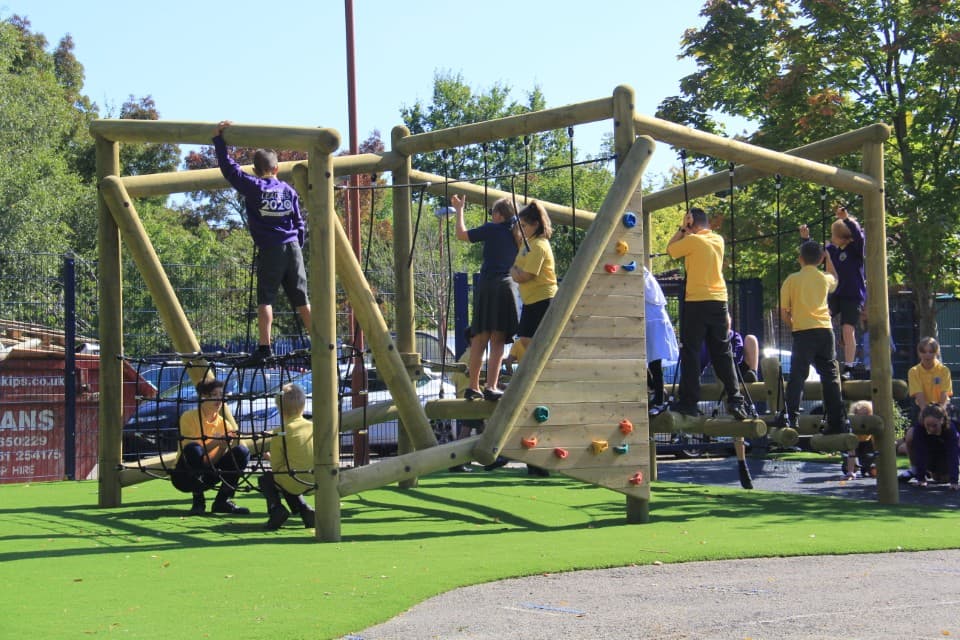 A group of children are climbing on a wooden climbing frame. The frame has a variety of different obstacles, encouraging the kids to take different paths to climb it.