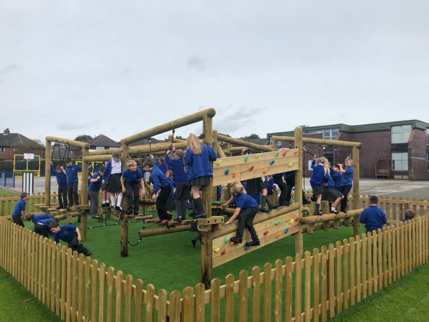 The Grizedale Forest Circuit installed on an artificial grass playground. The Forest Circuit has a variety of different challenges on it such as climbing, balancing, swinging and more. The play area is fenced with a small wooden fence. Children are playing all over the Forest Circuit.